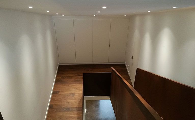 Escalier avec marches en bois et rampe brun foncé. Murs blancs, éclairage encastré et placards blancs intégrés sont visibles.