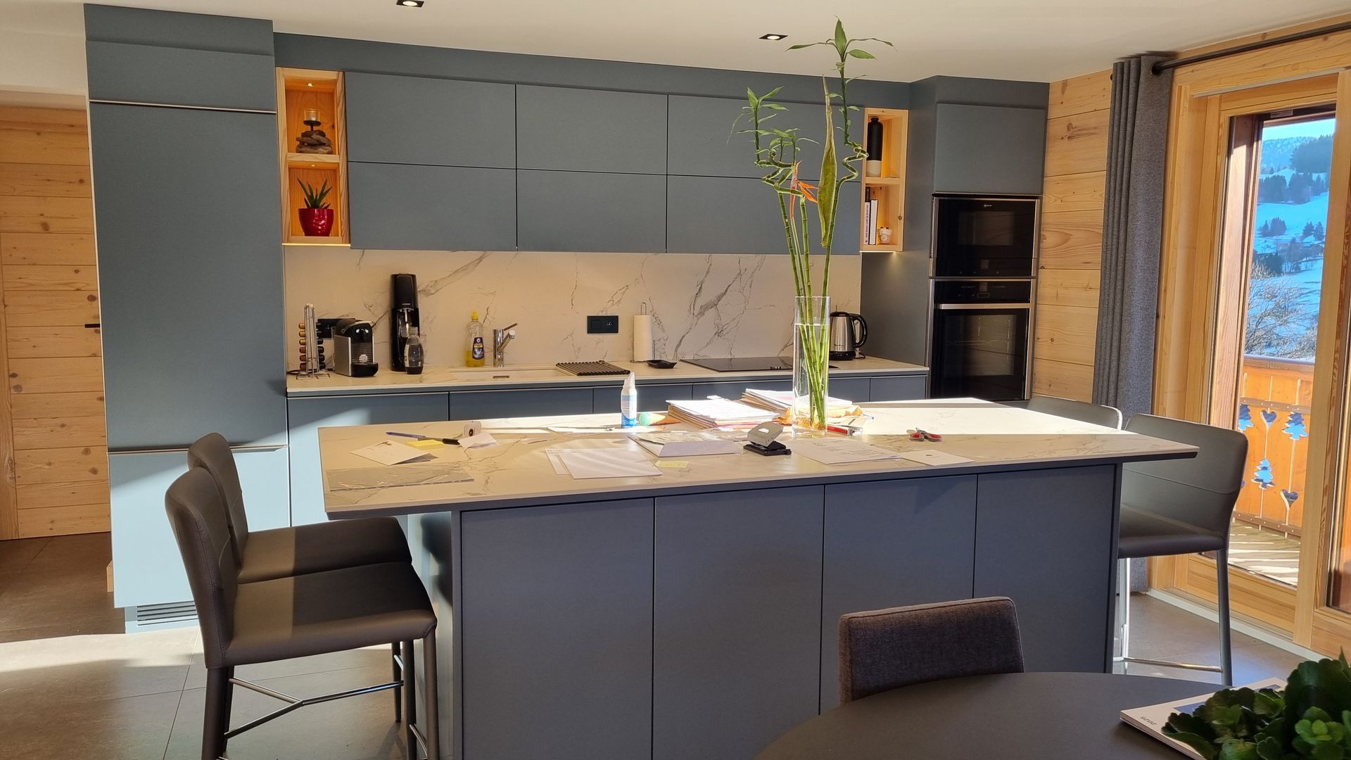 Cuisine moderne avec placards bleus, îlot central et tabourets de bar. Éléments en bois, électroménager et lumière naturelle.