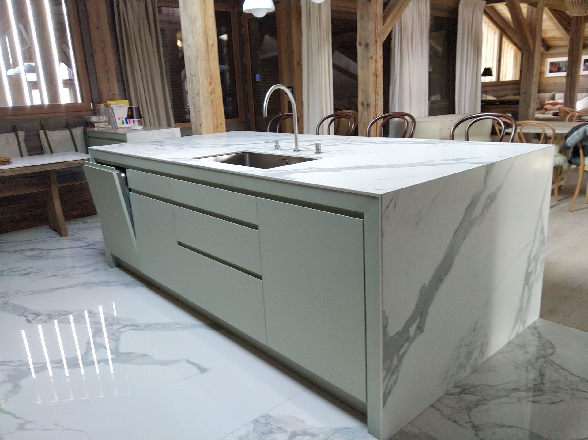 Îlot de cuisine avec comptoirs clairs, évier et robinet intégrés. Armoires vert pâle et sol clair. Poutres apparentes en bois rustique.