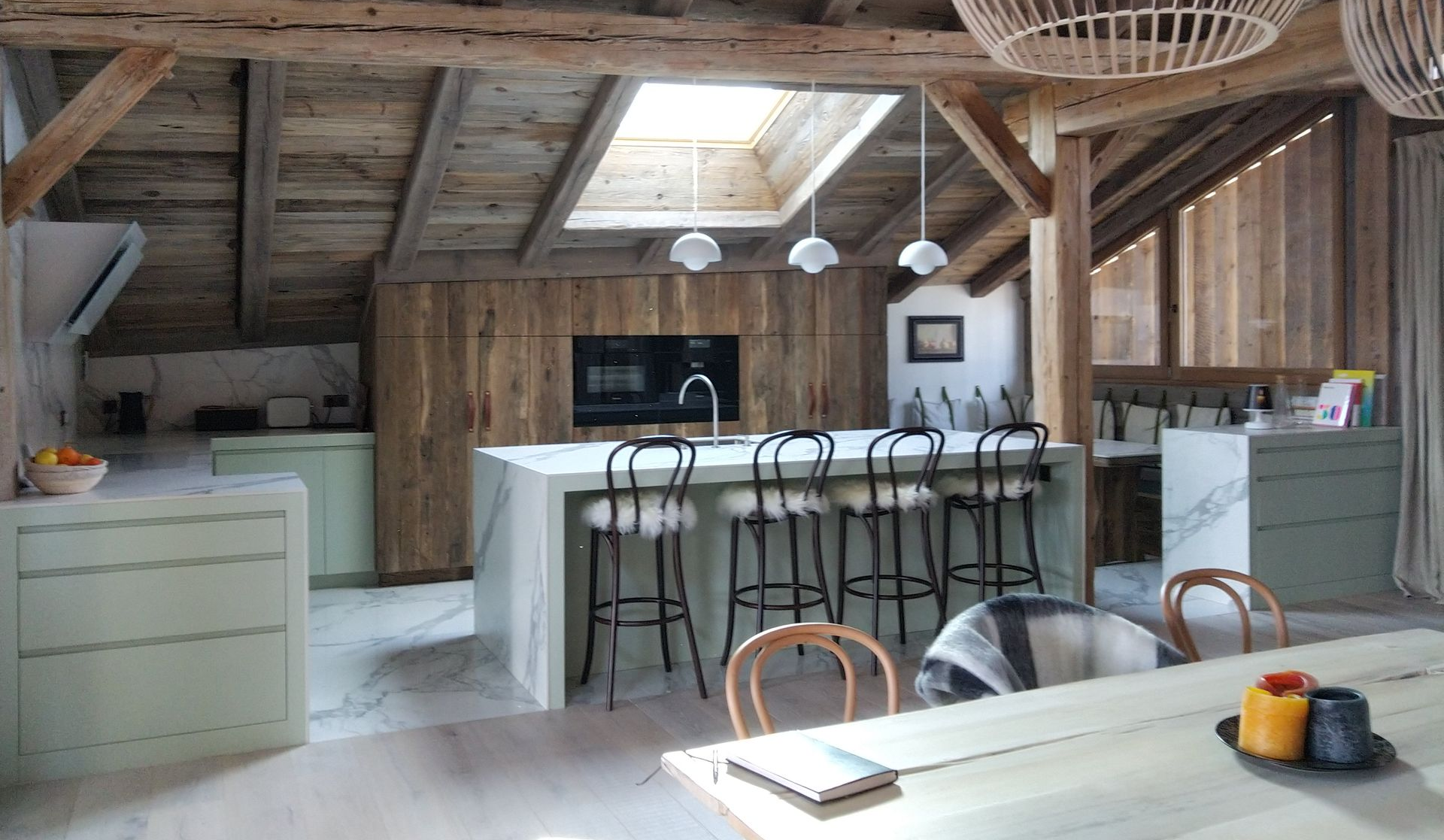 Cuisine rustique avec poutres en bois, îlot avec tabourets et puits de lumière.