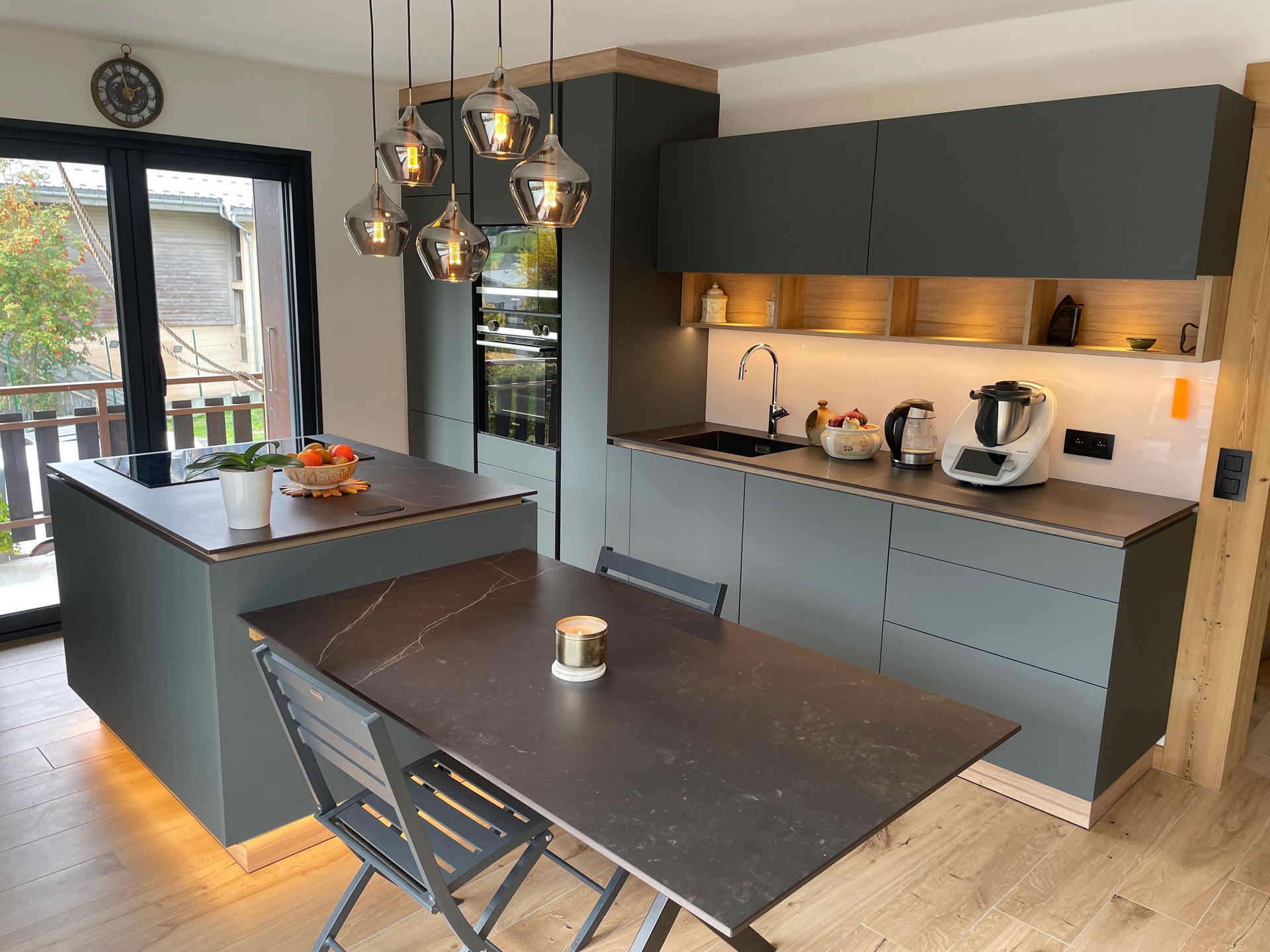 Cuisine moderne avec placards gris foncé, îlot central et table à manger, agrémentée de touches de bois et de suspensions lumineuses.