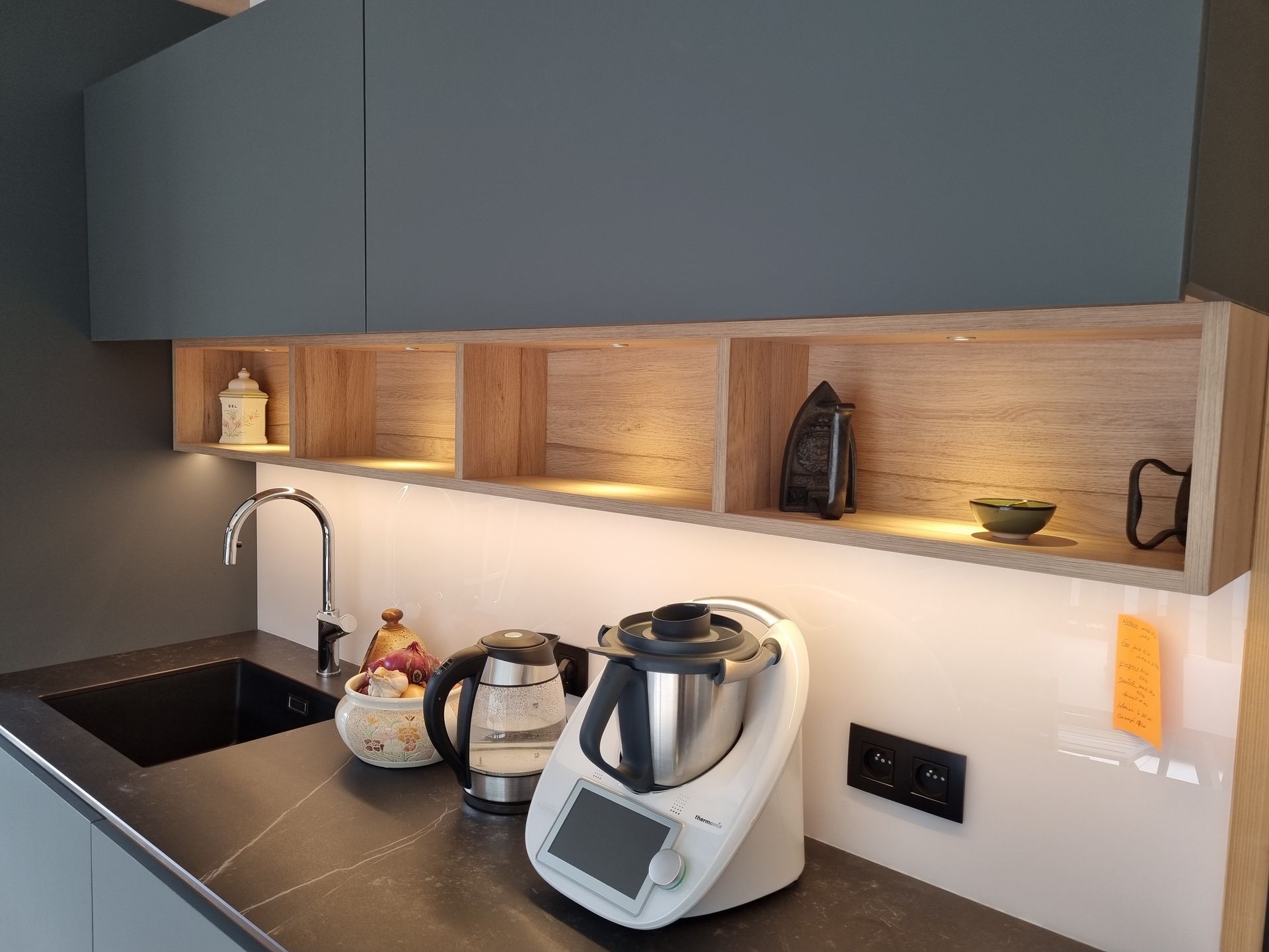 Plan de travail de cuisine avec évier, bouilloire, robot culinaire et étagère en bois encastrée éclairée par des spots sous les meubles.