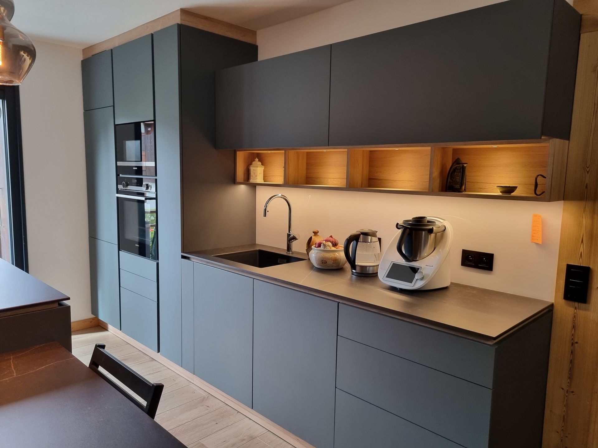 Cuisine moderne avec armoires gris foncé, électroménagers intégrés, évier en acier inoxydable et éléments en bois.