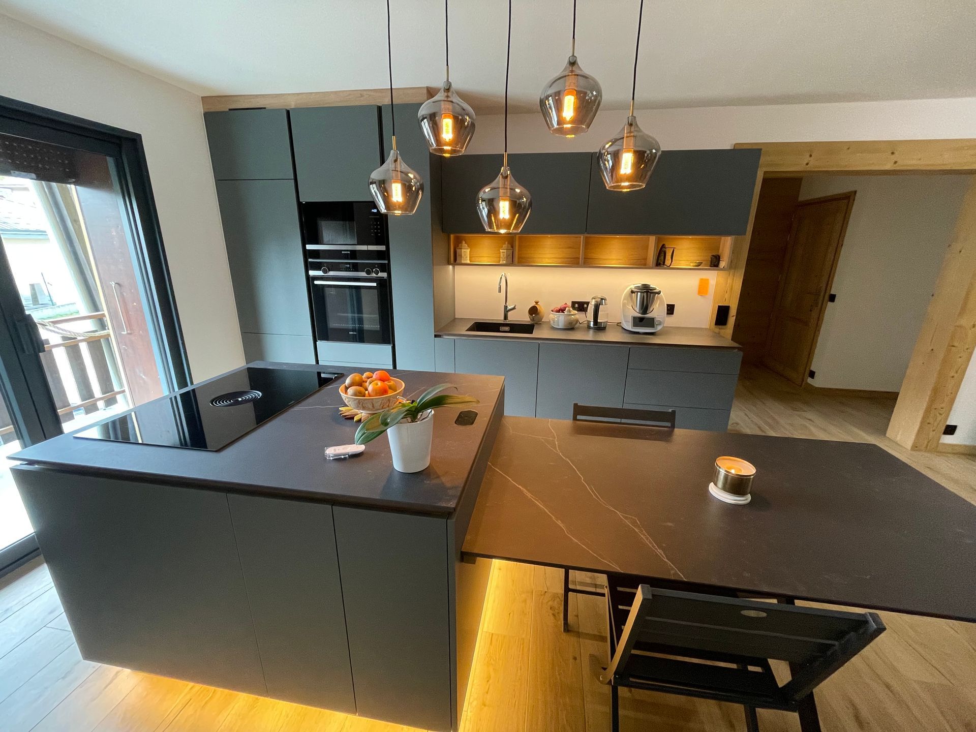 Cuisine moderne avec placards gris, îlot central avec table extensible et luminaires suspendus.