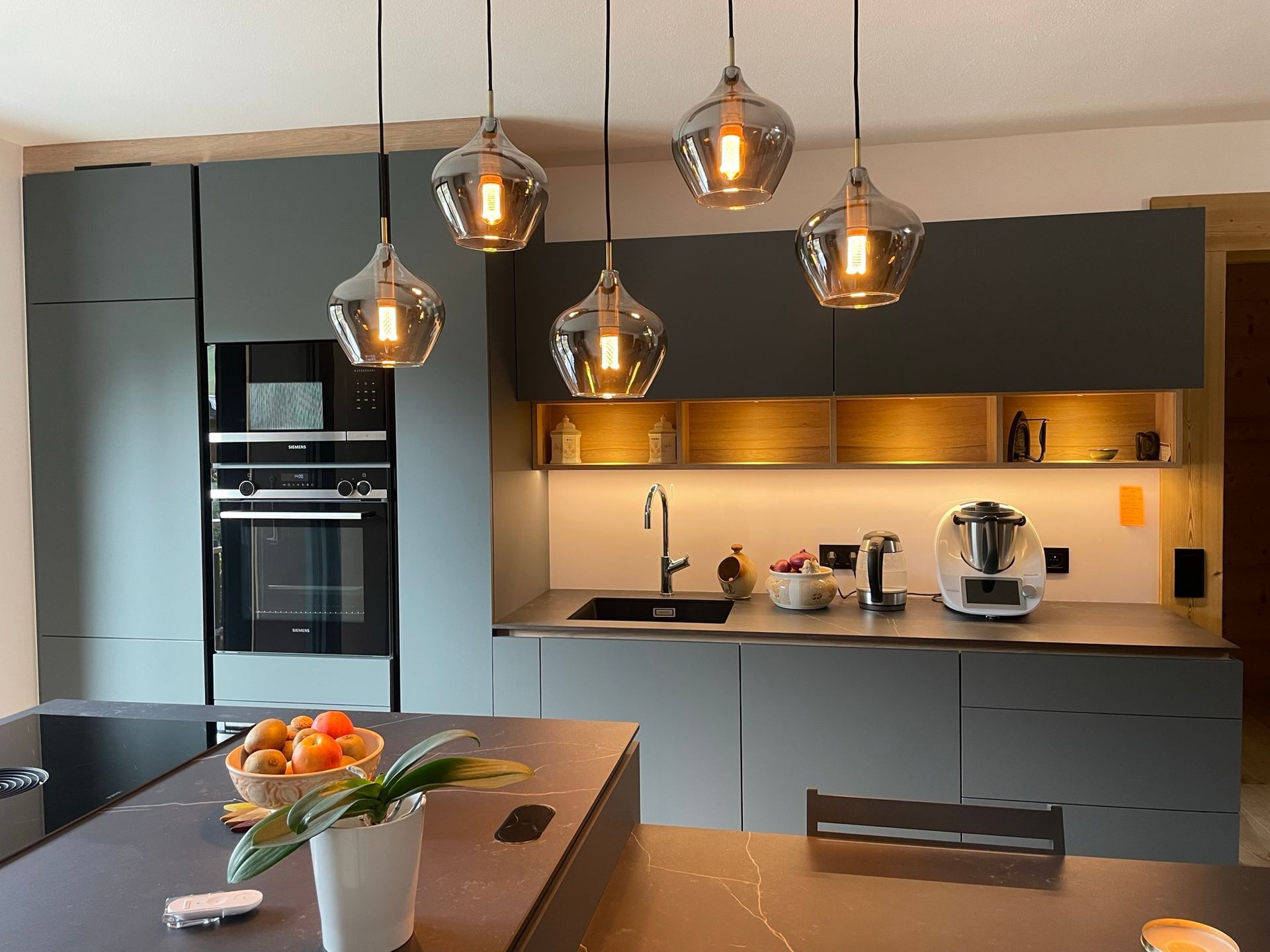 Cuisine moderne avec placards gris, îlot central et suspensions lumineuses. On aperçoit également un four encastré, un évier et des appareils électroménagers posés sur le plan de travail.