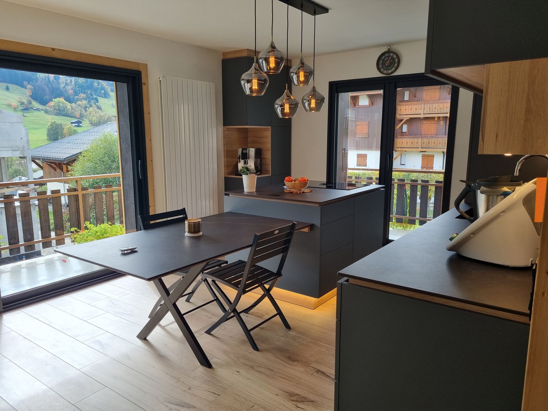 Cuisine moderne avec îlot central, table et vue sur la montagne. Placards gris foncé, éléments en bois, suspensions lumineuses.