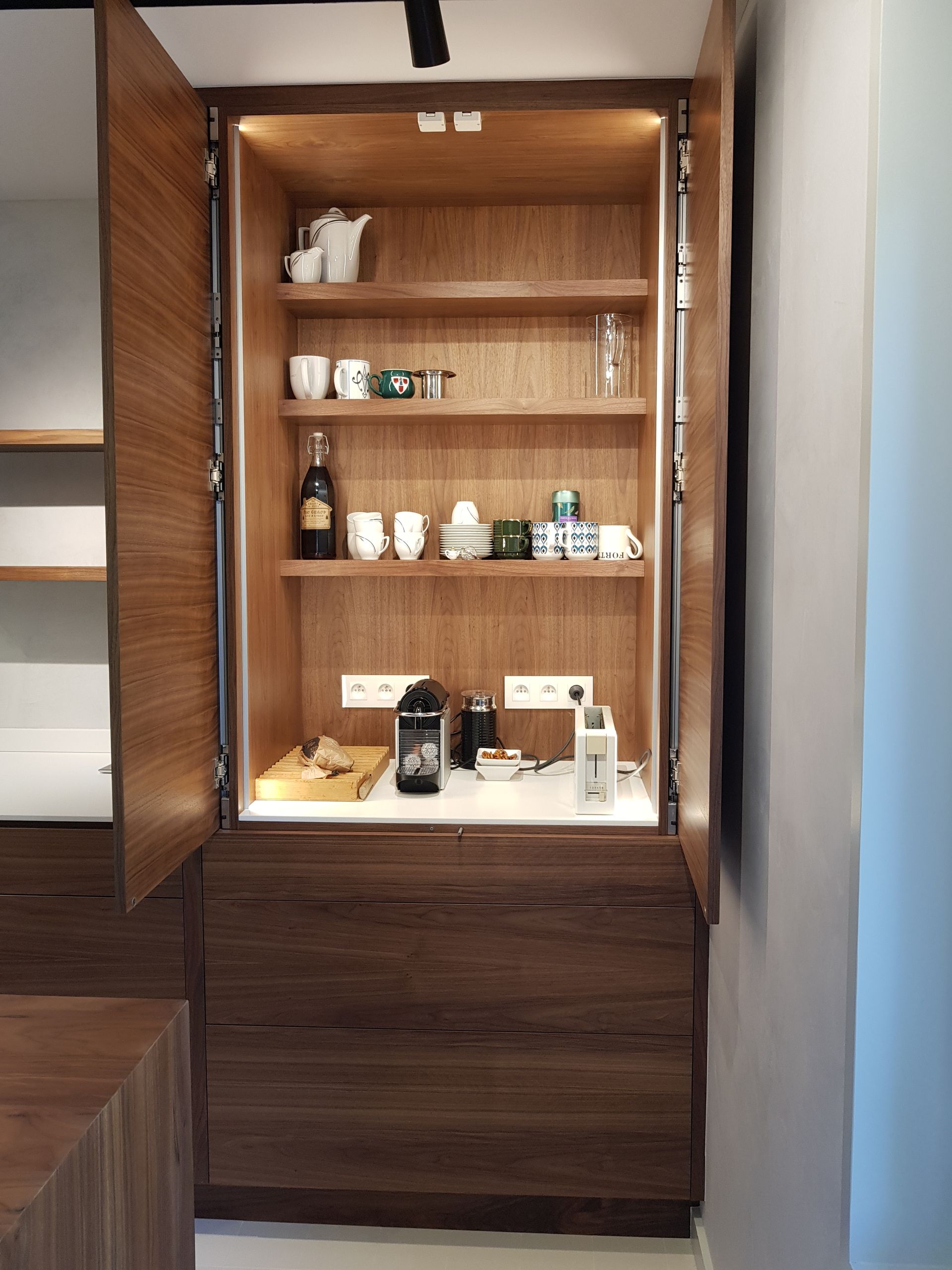 Un meuble en bois s'ouvre, révélant des étagères garnies d'accessoires pour la préparation du café et de tasses, éclairées par des lumières.