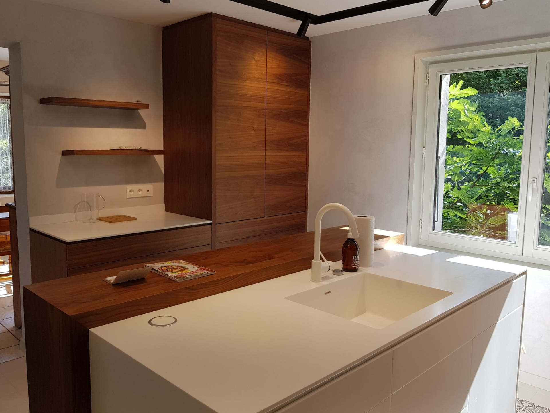 Cuisine moderne avec comptoirs blancs, armoires en bois et grande fenêtre.