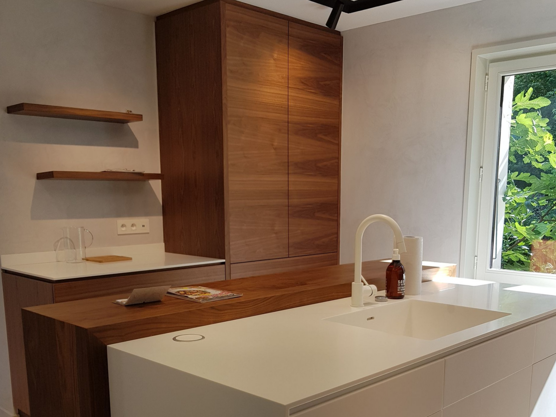 Cuisine moderne avec placards en bois, plan de travail blanc, évier et fenêtre ouverte.
