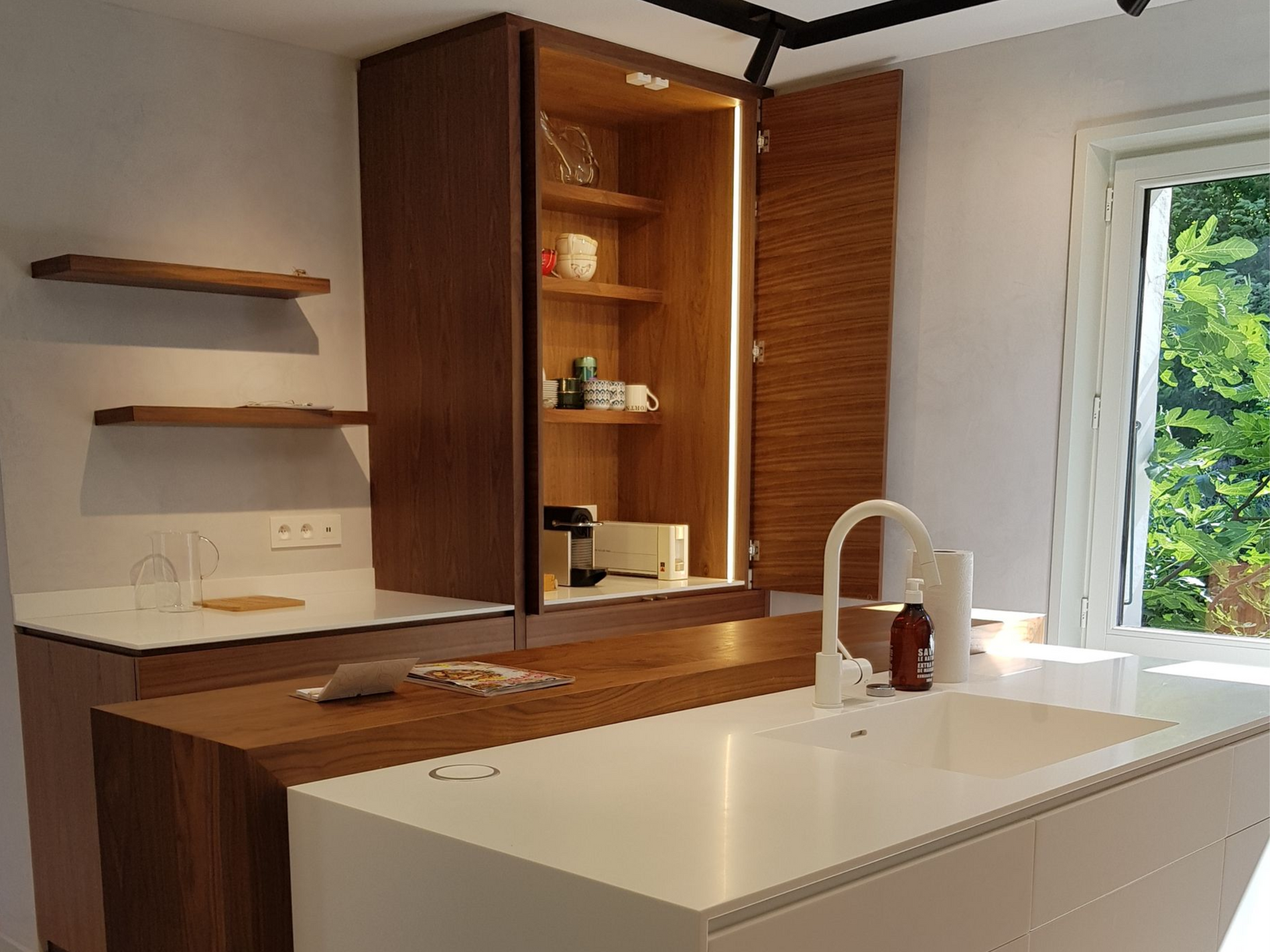 Cuisine avec placards en bois, plan de travail blanc et évier. La lumière du soleil entre par une fenêtre.