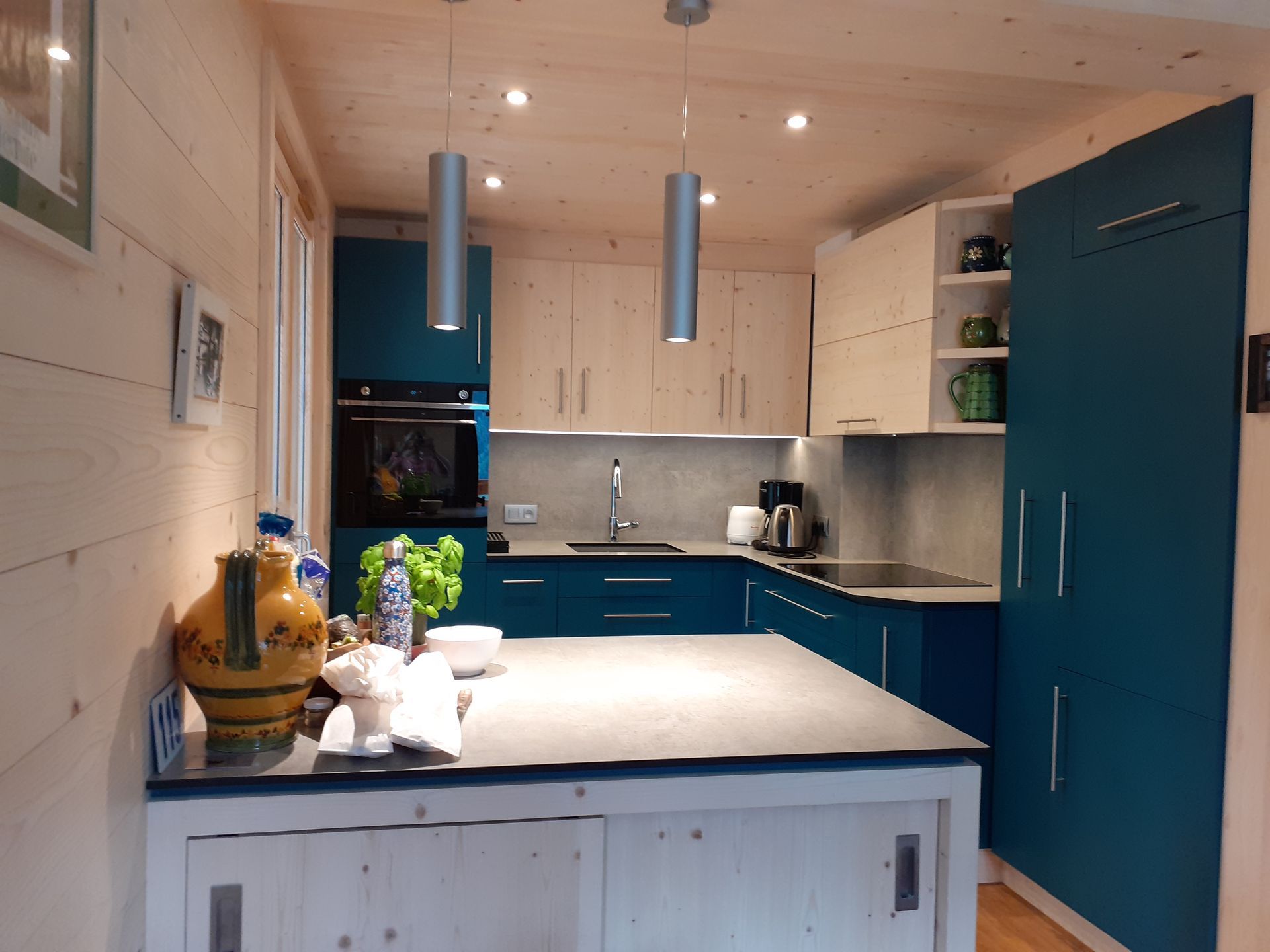 Cuisine avec placards bleu sarcelle, murs en bois clair et plan de travail gris. Deux suspensions argentées éclairent le plafond.