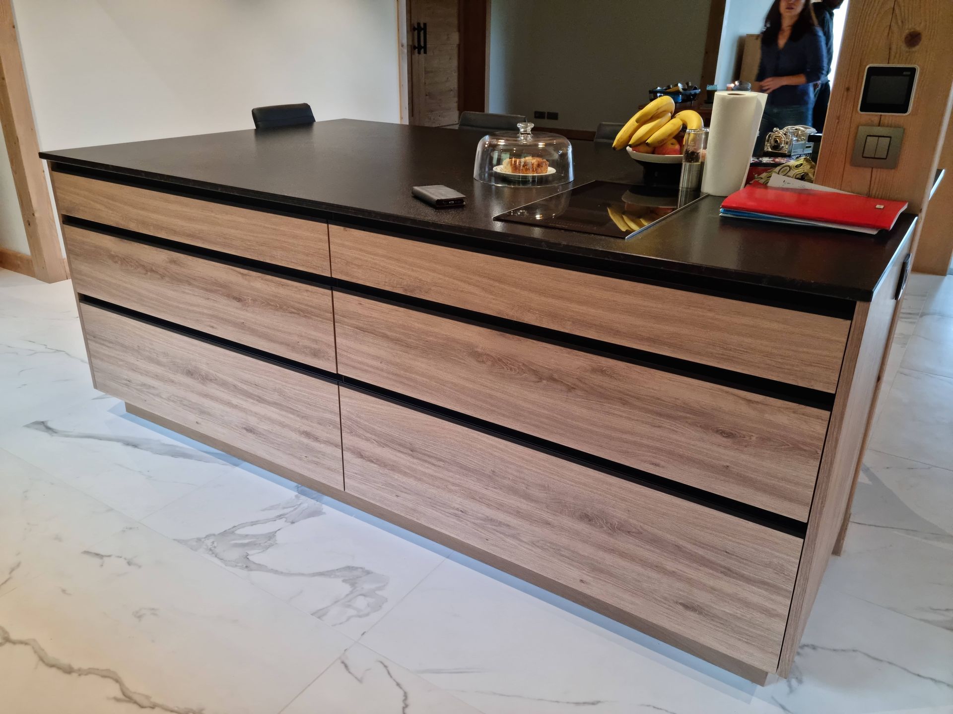 Îlot de cuisine avec comptoir noir, armoires en bois et moulures noires. Une personne se tient à proximité.
