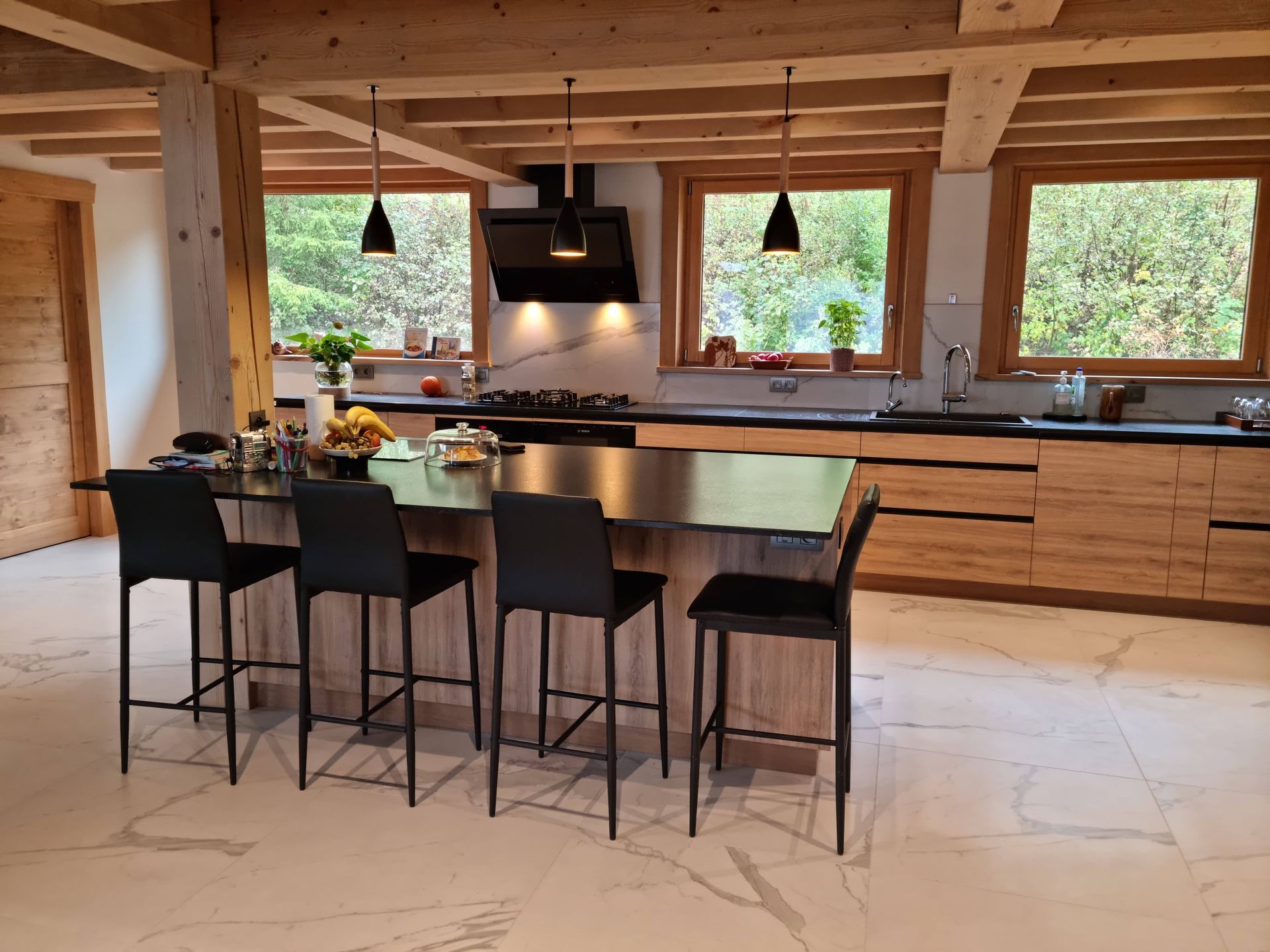 Cuisine moderne avec poutres apparentes, îlot central avec tabourets noirs, fenêtres donnant sur la verdure.