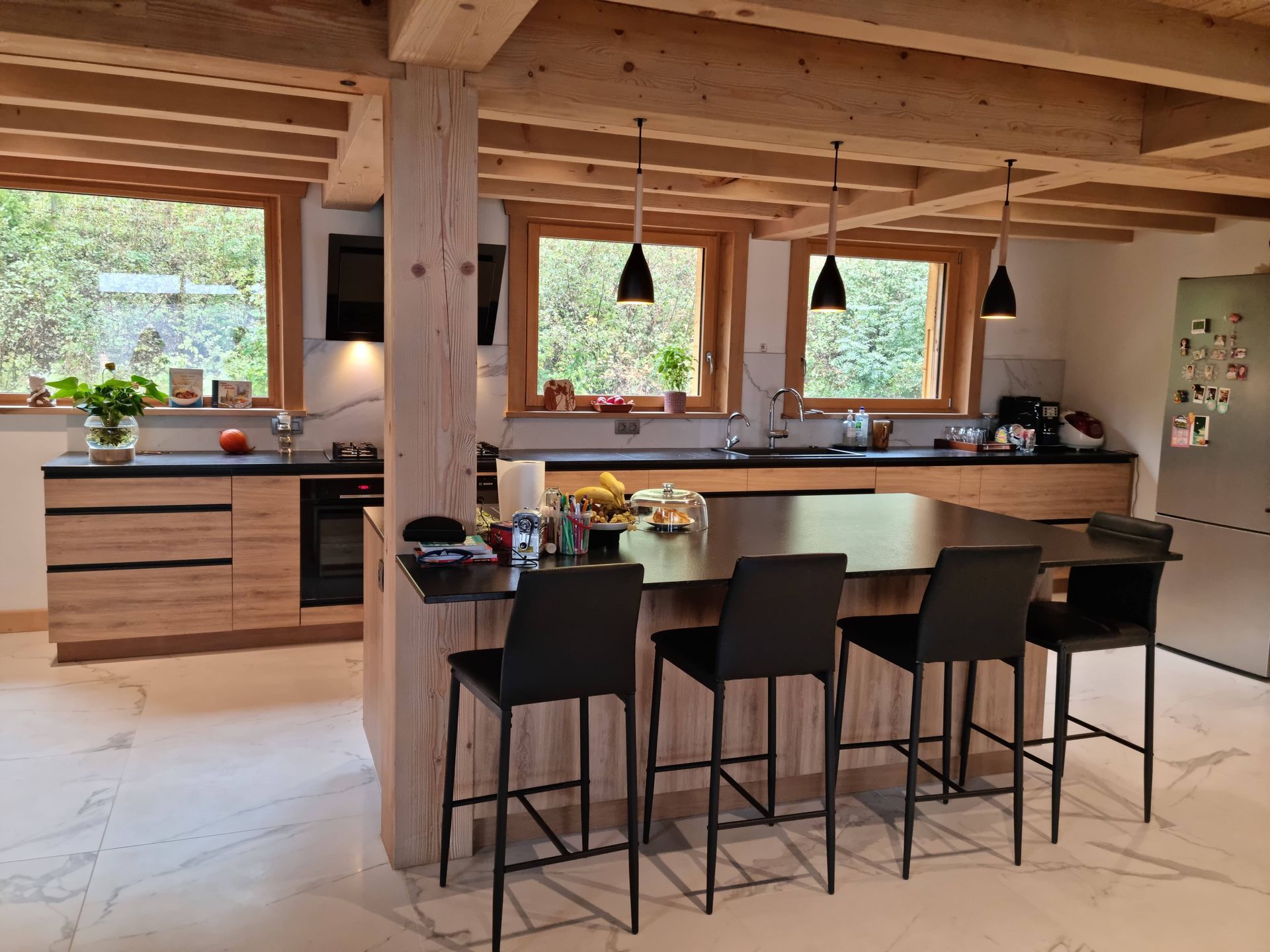 Cuisine moderne avec des meubles en bois clair, un îlot central sombre avec des tabourets noirs et des poutres apparentes.