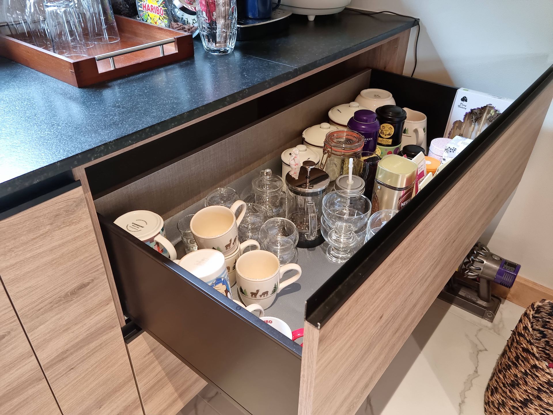 Tiroir de cuisine ouvert, rempli de tasses, de verres et de récipients. Le tiroir se trouve à l'intérieur d'un meuble en bois.