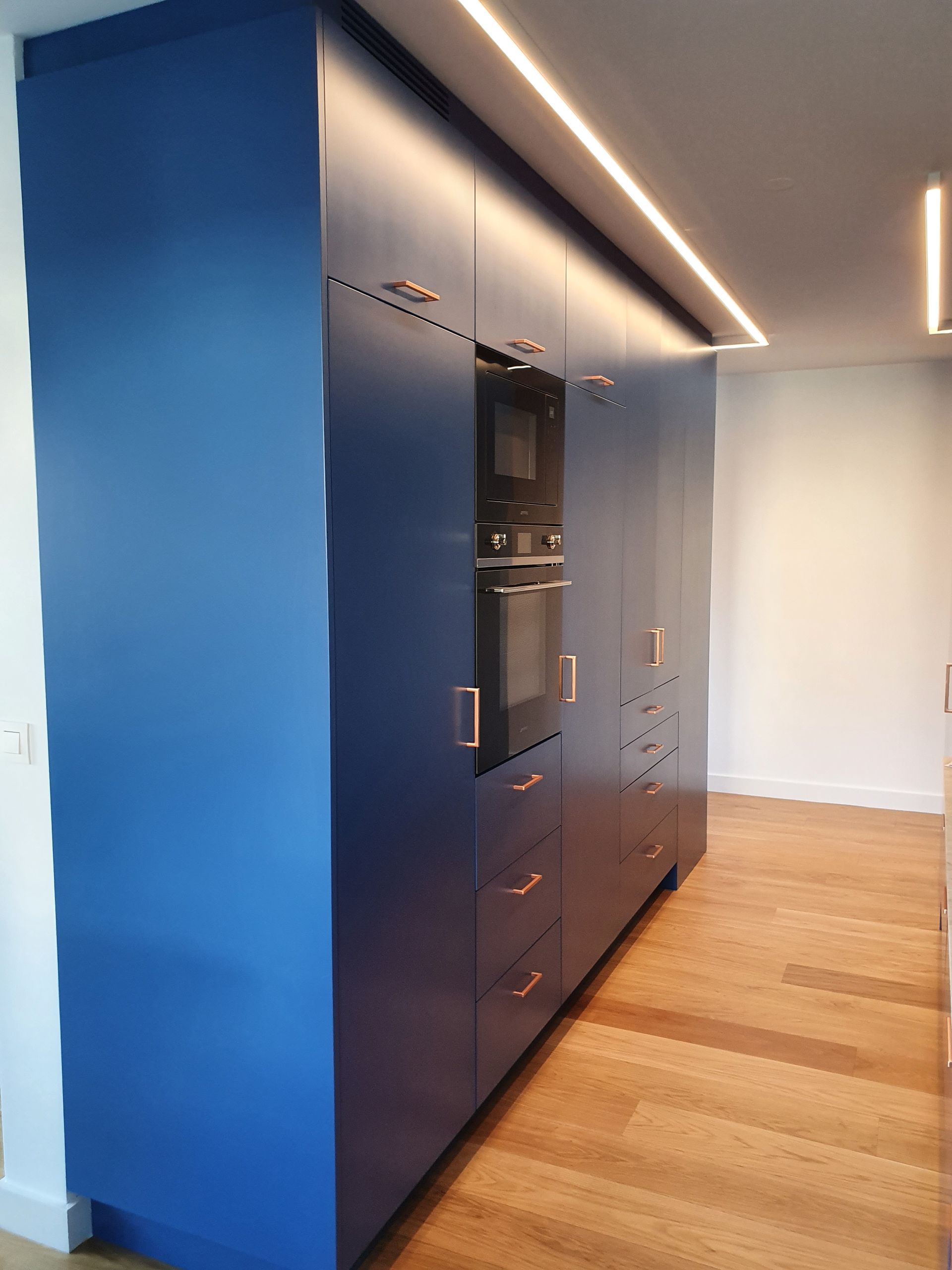 Cuisine équipée d'armoires bleues avec poignées en cuivre, four encastré et parquet. Éclairage encastré.