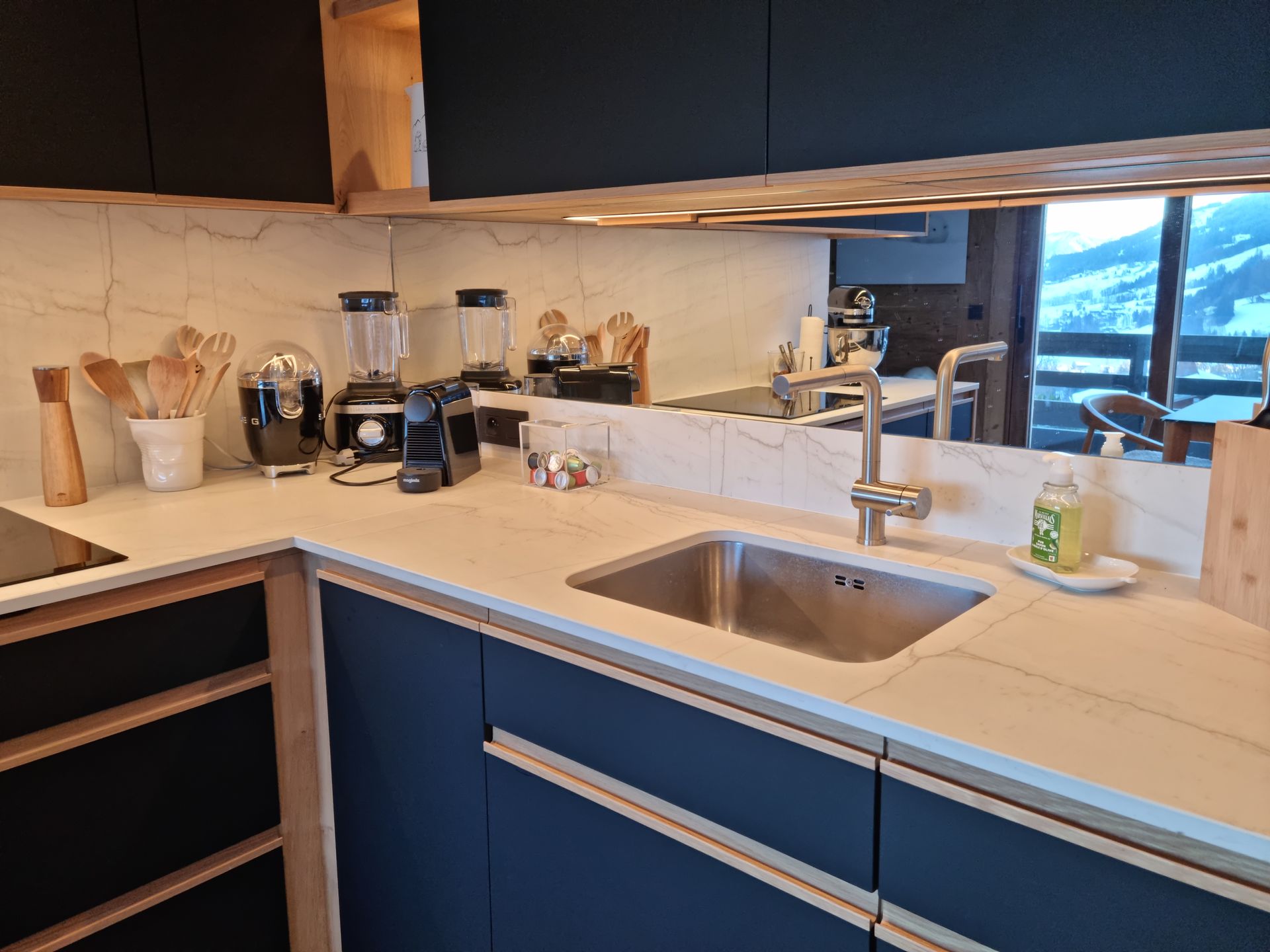 Cuisine moderne avec armoires noir foncé, comptoir clair, évier en acier inoxydable et un miroir reflétant une vue sur la montagne.