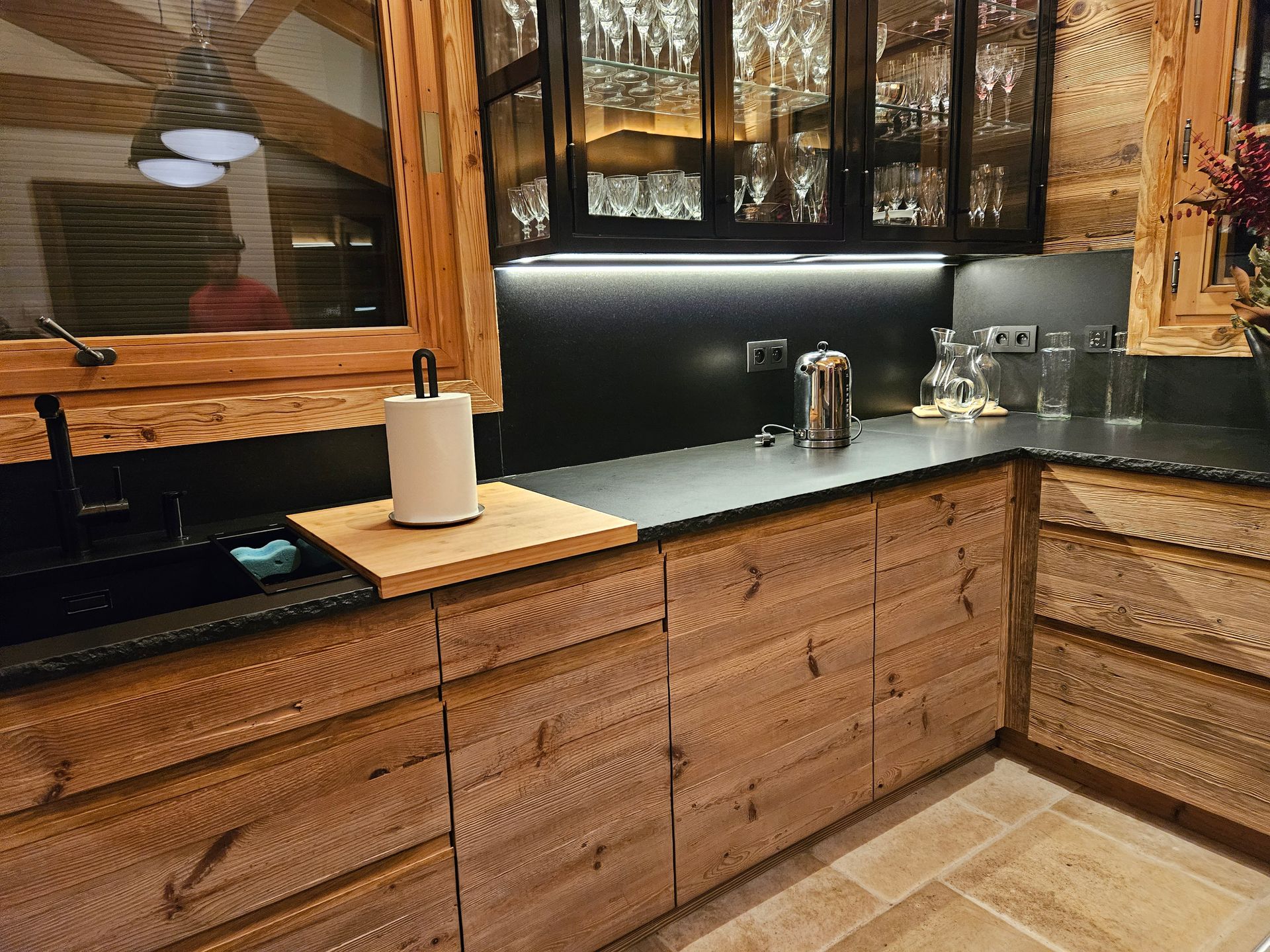 Cuisine rustique avec placards en bois, plan de travail noir, évier et fenêtre.