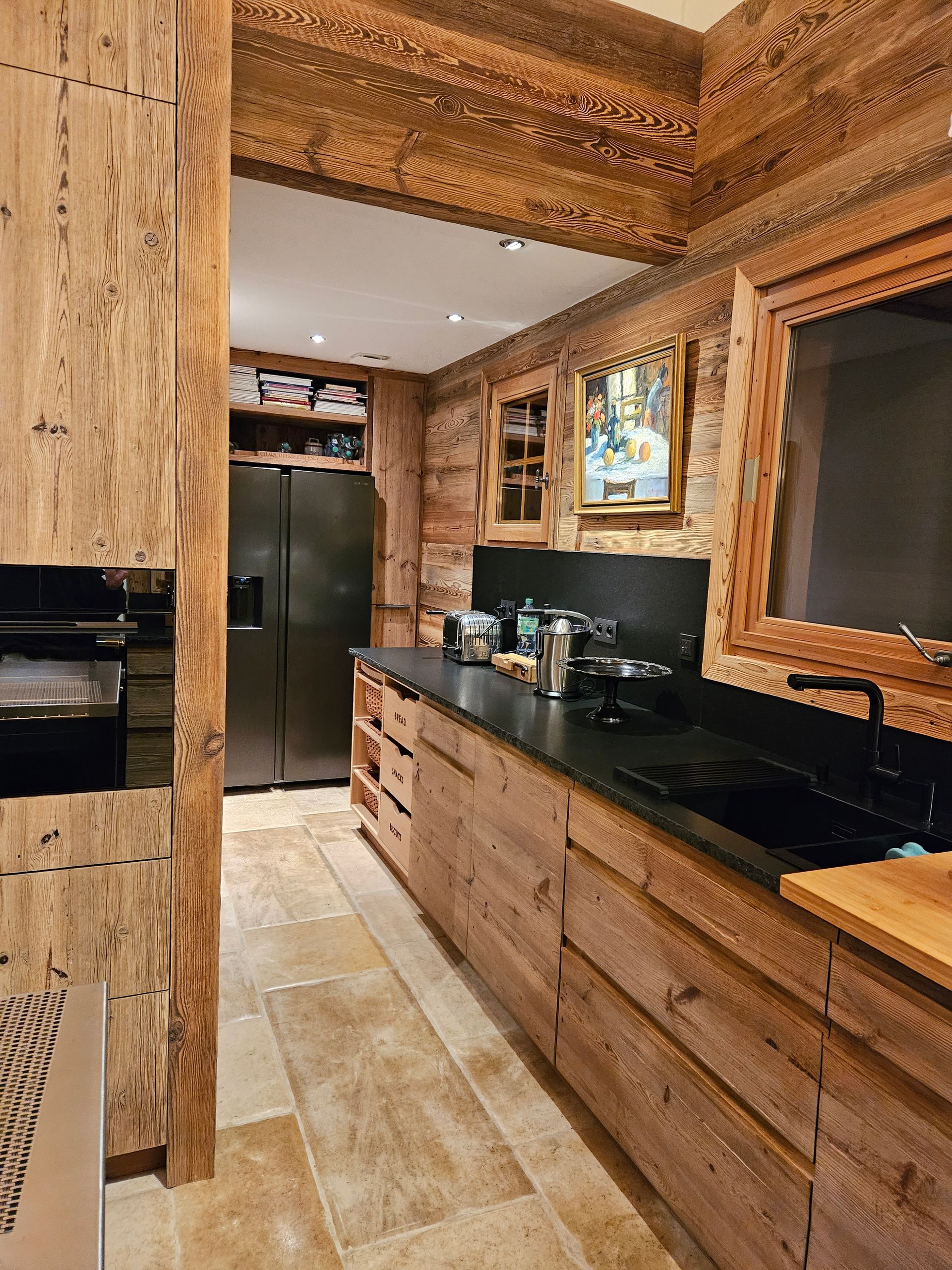 Cuisine en bois avec plans de travail noirs, réfrigérateur et sol en pierre.