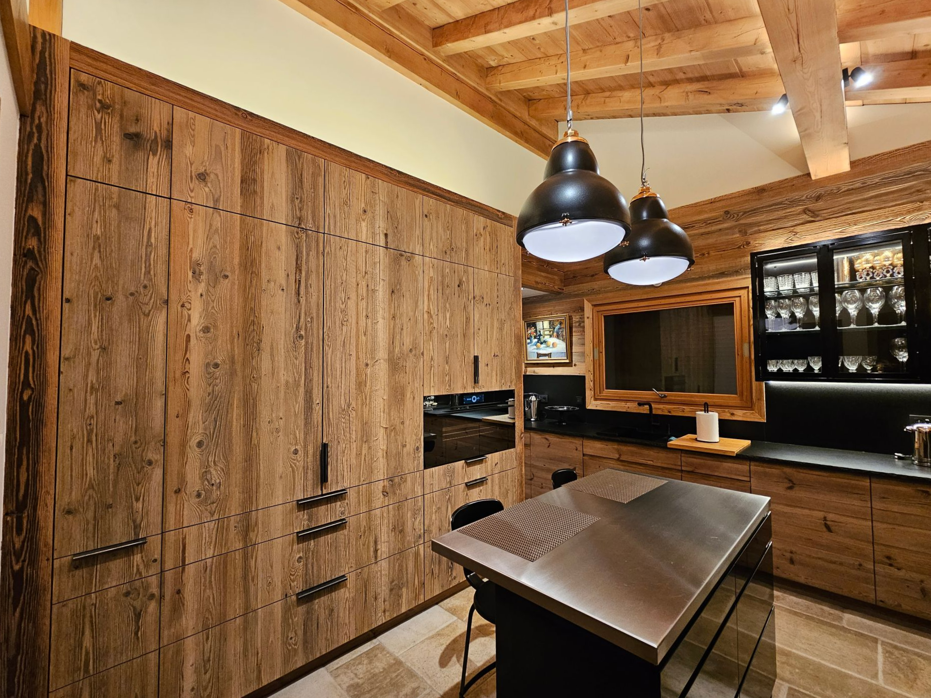 Cuisine rustique avec placards en bois et îlot central en acier inoxydable, éclairée par des suspensions lumineuses.