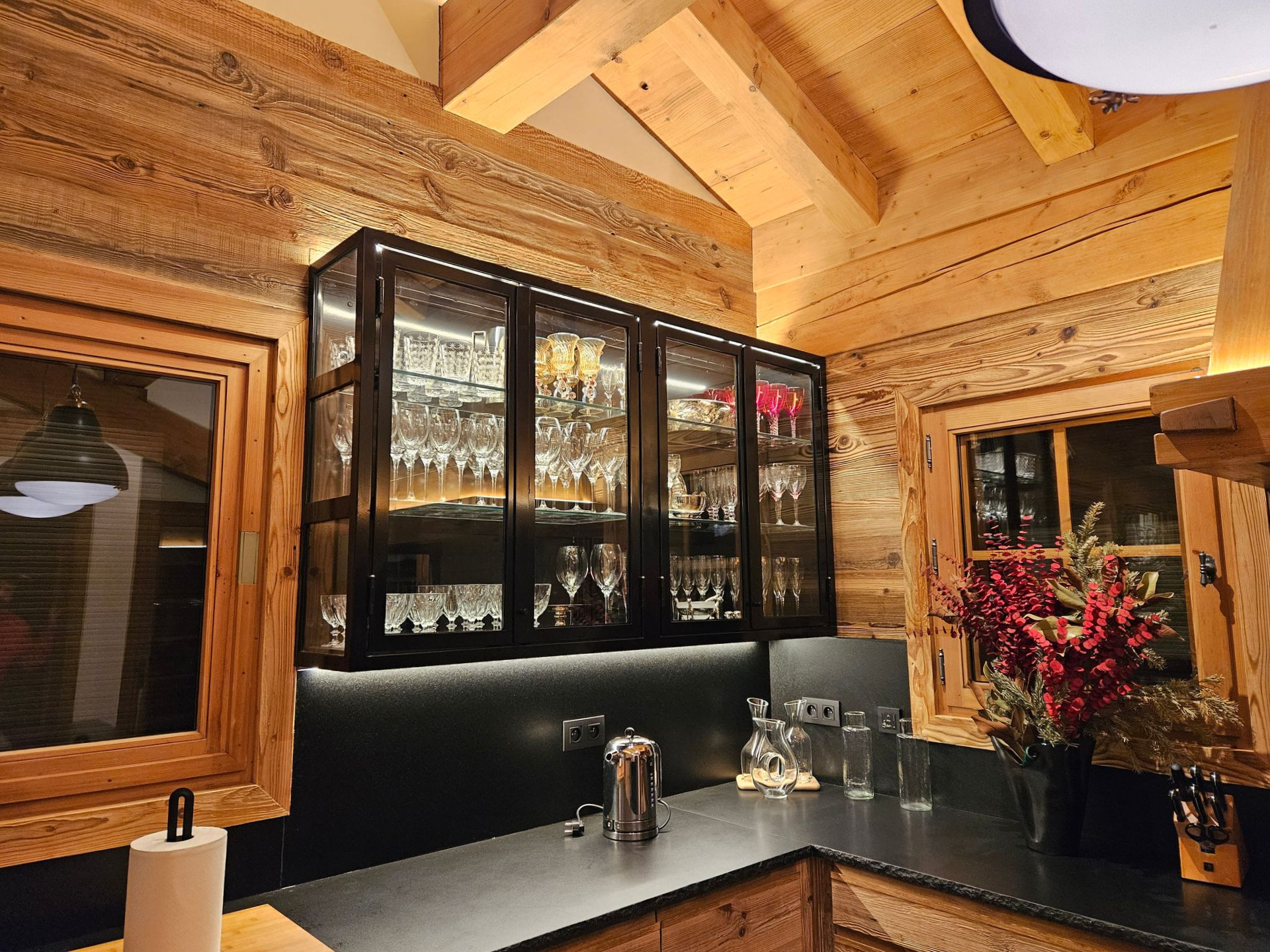 Cuisine lambrissée avec un plan de travail foncé et une vitrine remplie de verrerie.