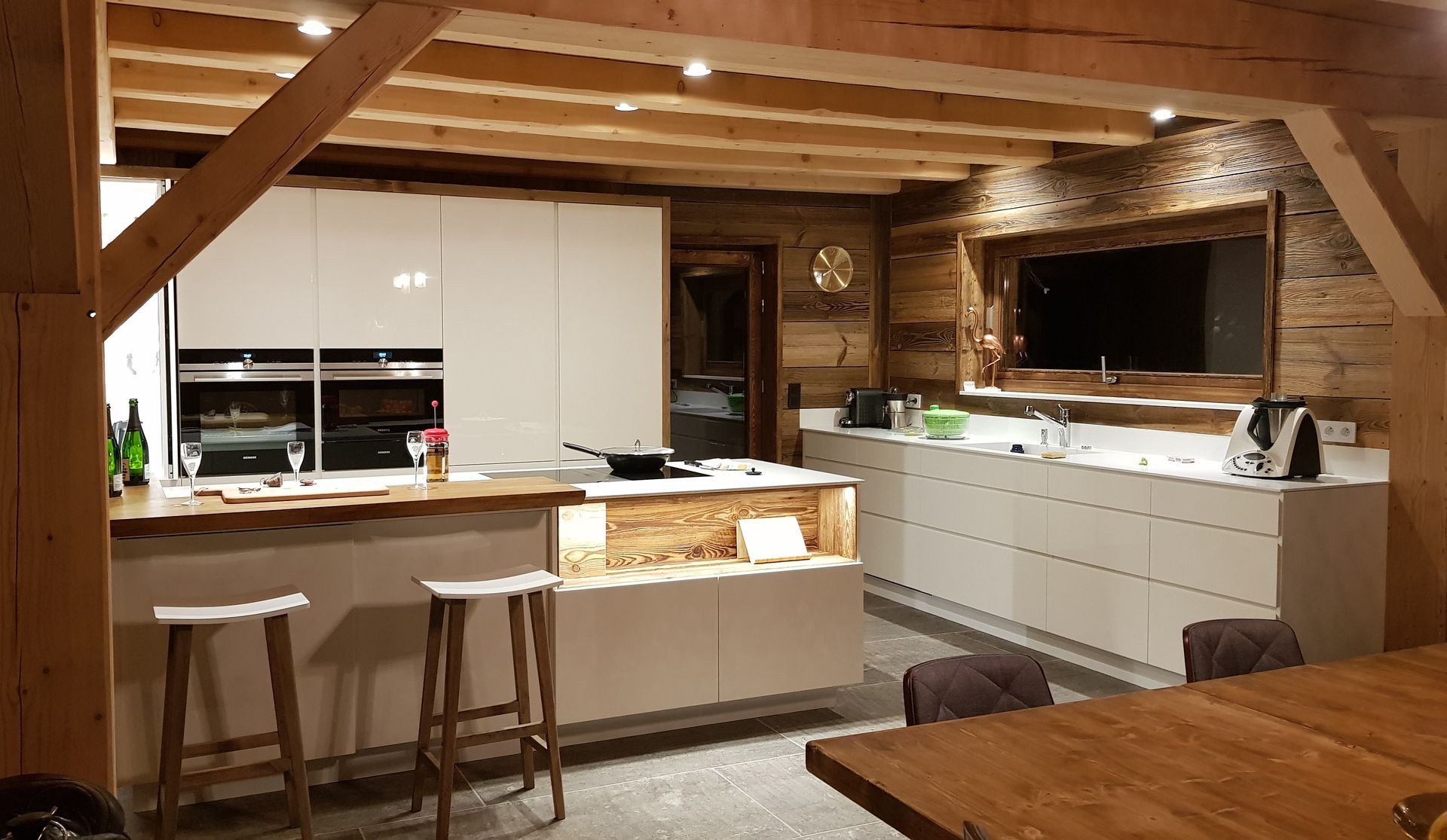 Cuisine moderne avec poutres et lambris en bois, armoires blanches et îlot avec tabourets de bar.