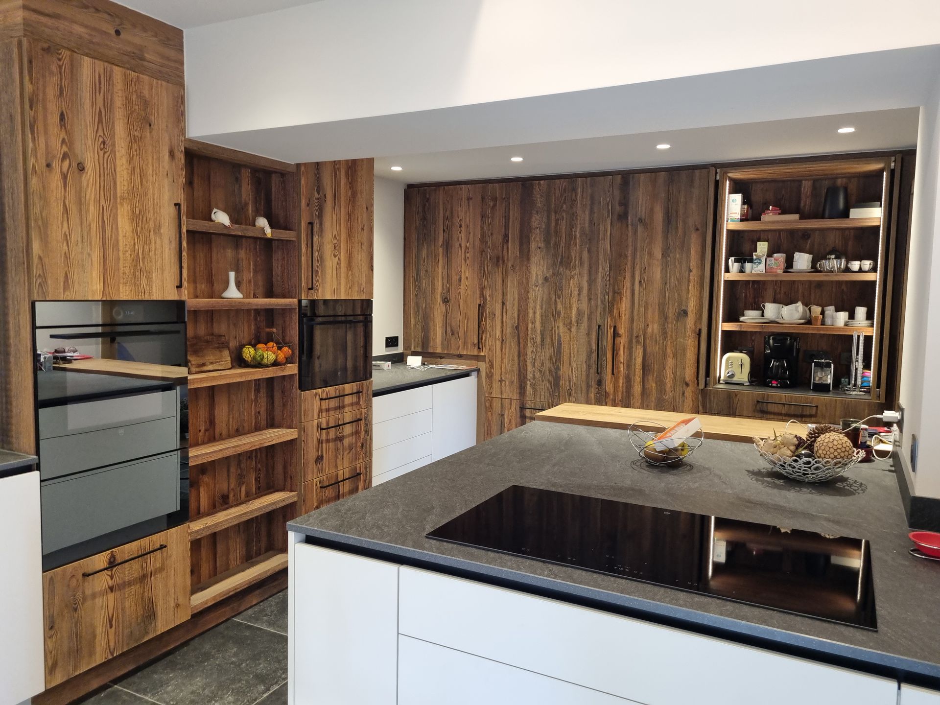 Cuisine rustique avec placards et îlot en bois, comptoirs noirs et appareils électroménagers intégrés.