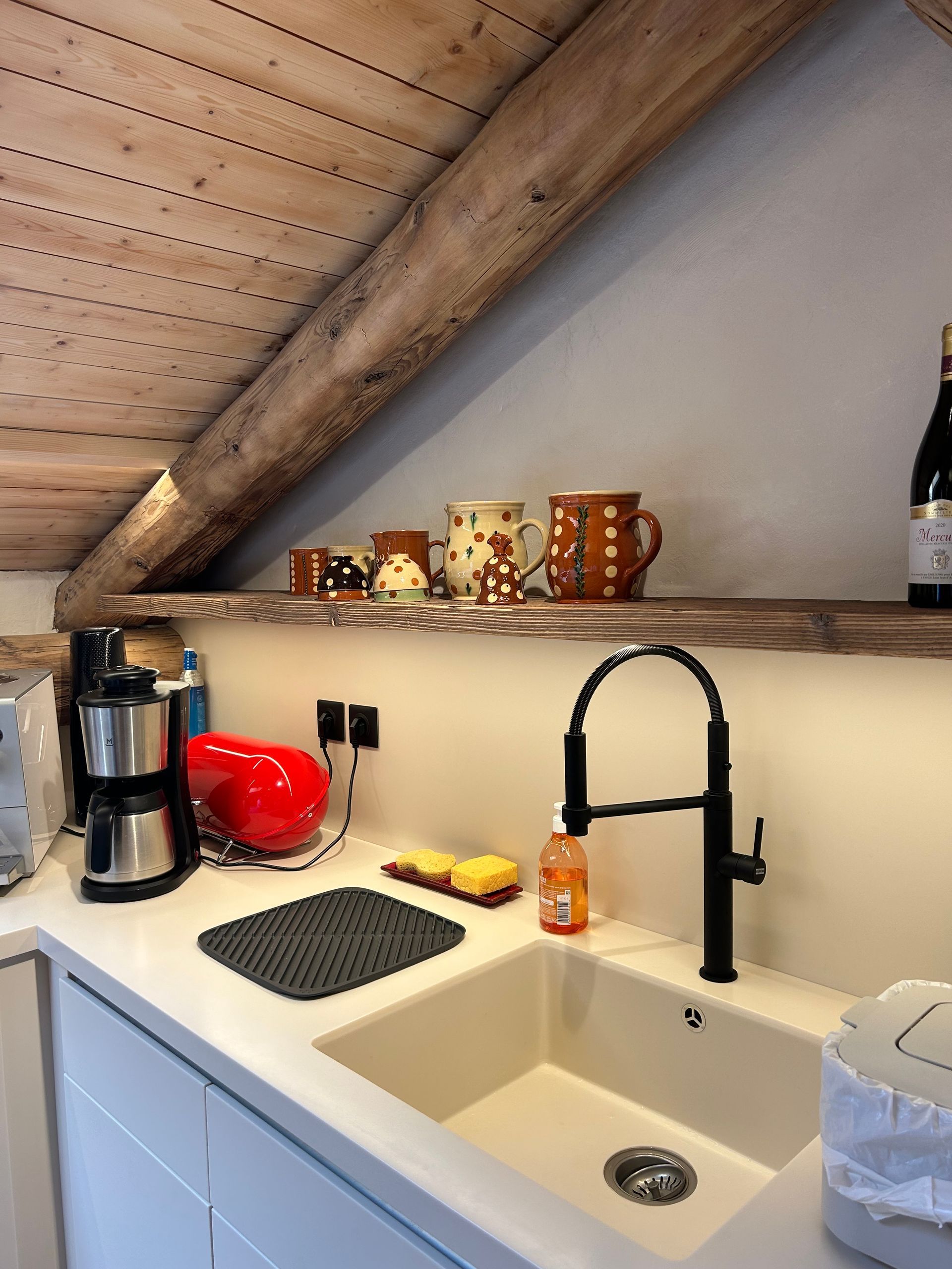 Cuisine avec évier, robinet, plan de travail, étagère avec tasses et plafond à poutres apparentes.