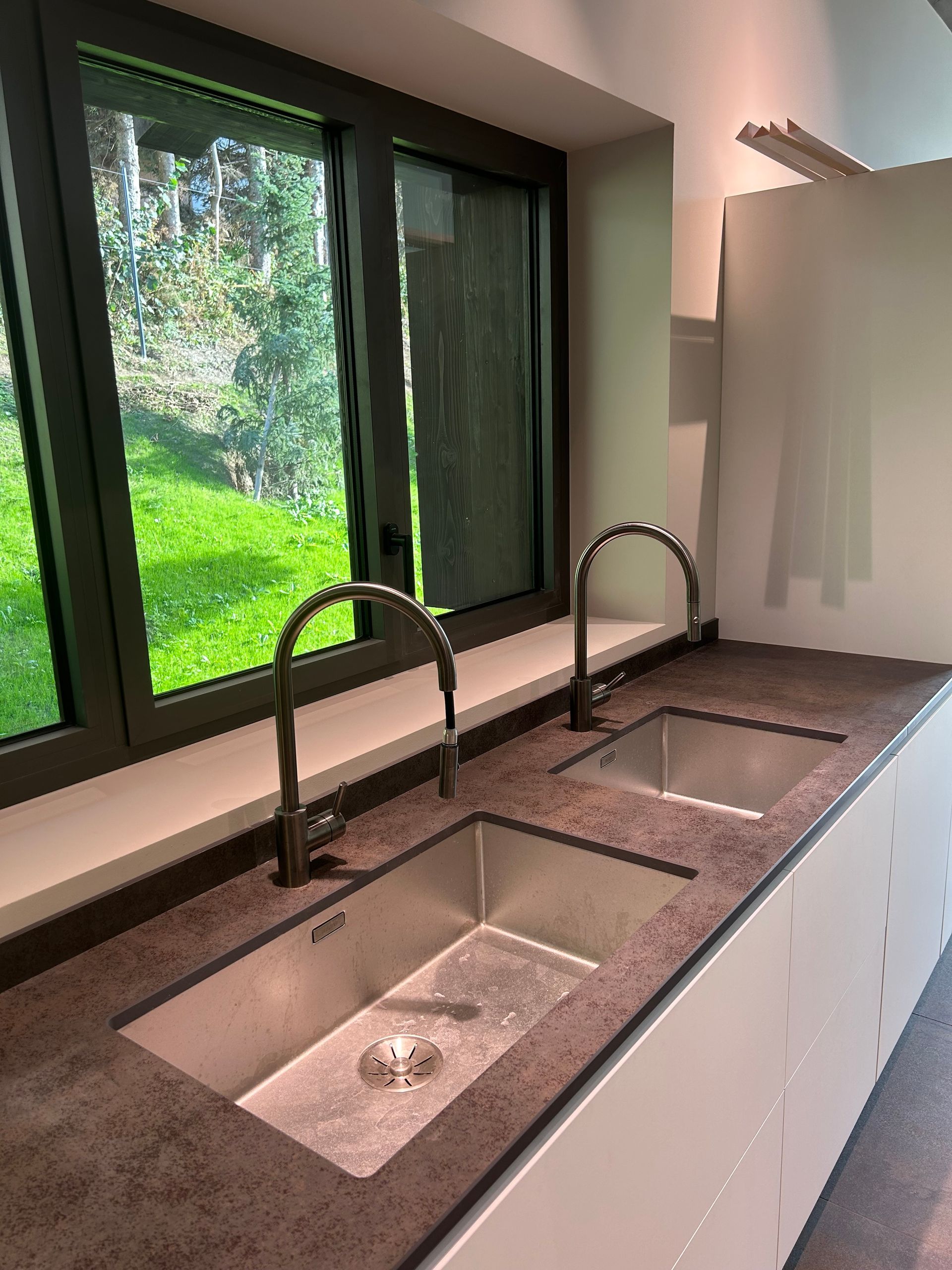 Deux éviers en acier inoxydable avec robinets arqués sur un plan de travail marron, à côté d'une fenêtre donnant sur une pelouse verte.