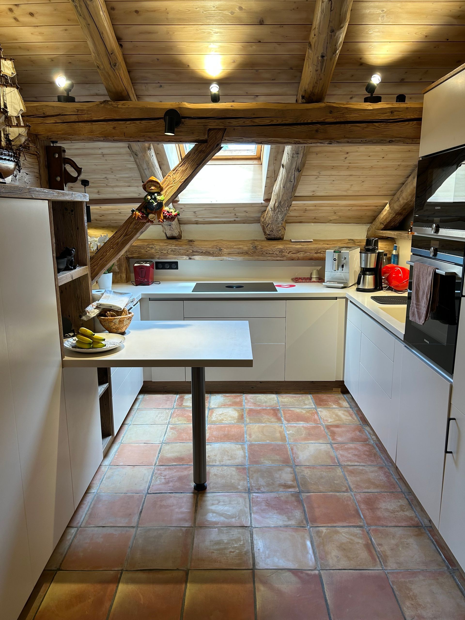 Cuisine avec poutres apparentes, placards blancs et sol en carrelage de terre cuite ; la table est attenante aux placards.