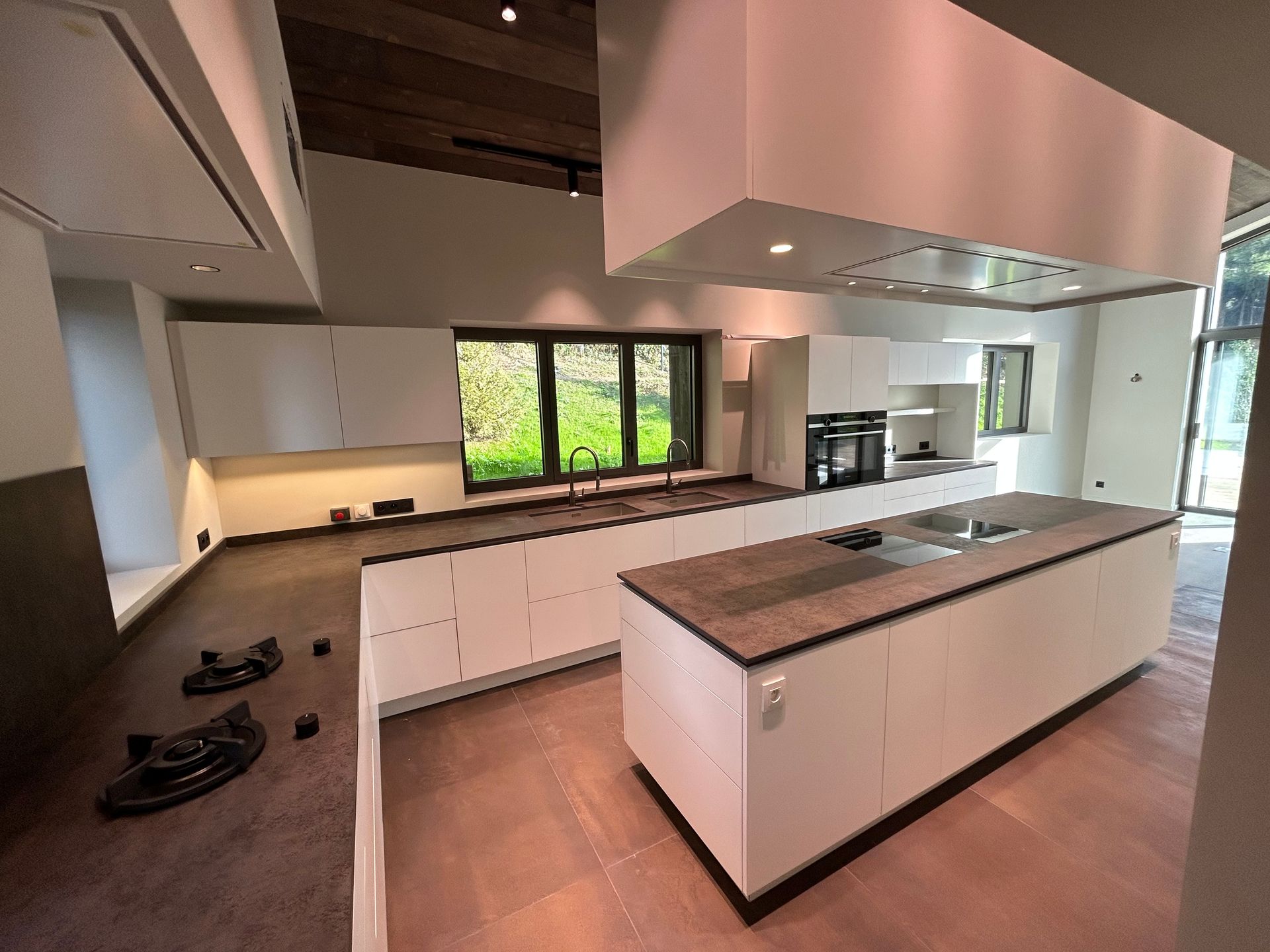 Cuisine moderne blanche avec îlot central, plans de travail foncés et grande fenêtre donnant sur la verdure.