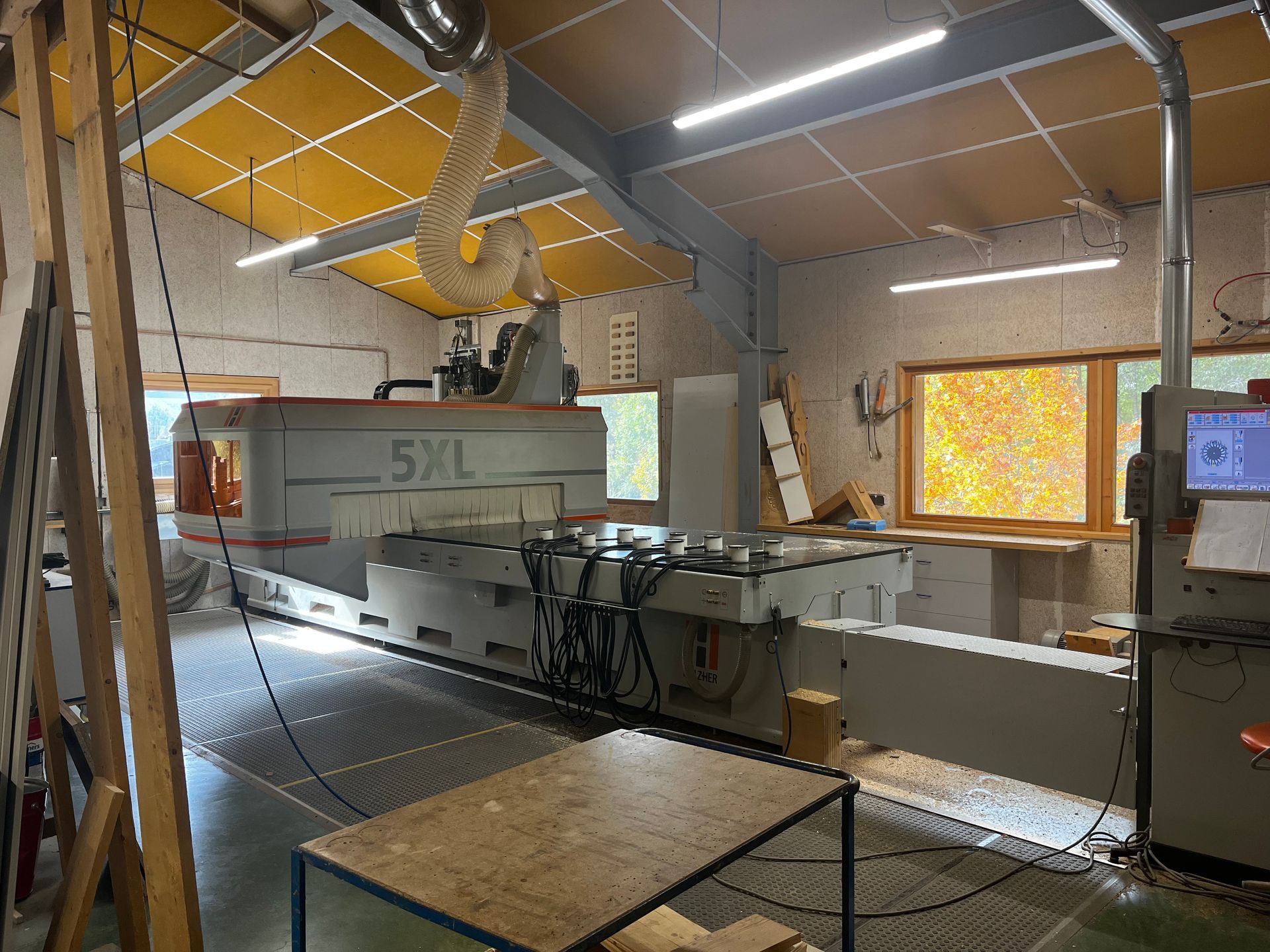 Une grande machine CNC portant l'inscription « 5XL » se trouve dans un atelier de menuiserie. La lumière du soleil entre par une fenêtre.