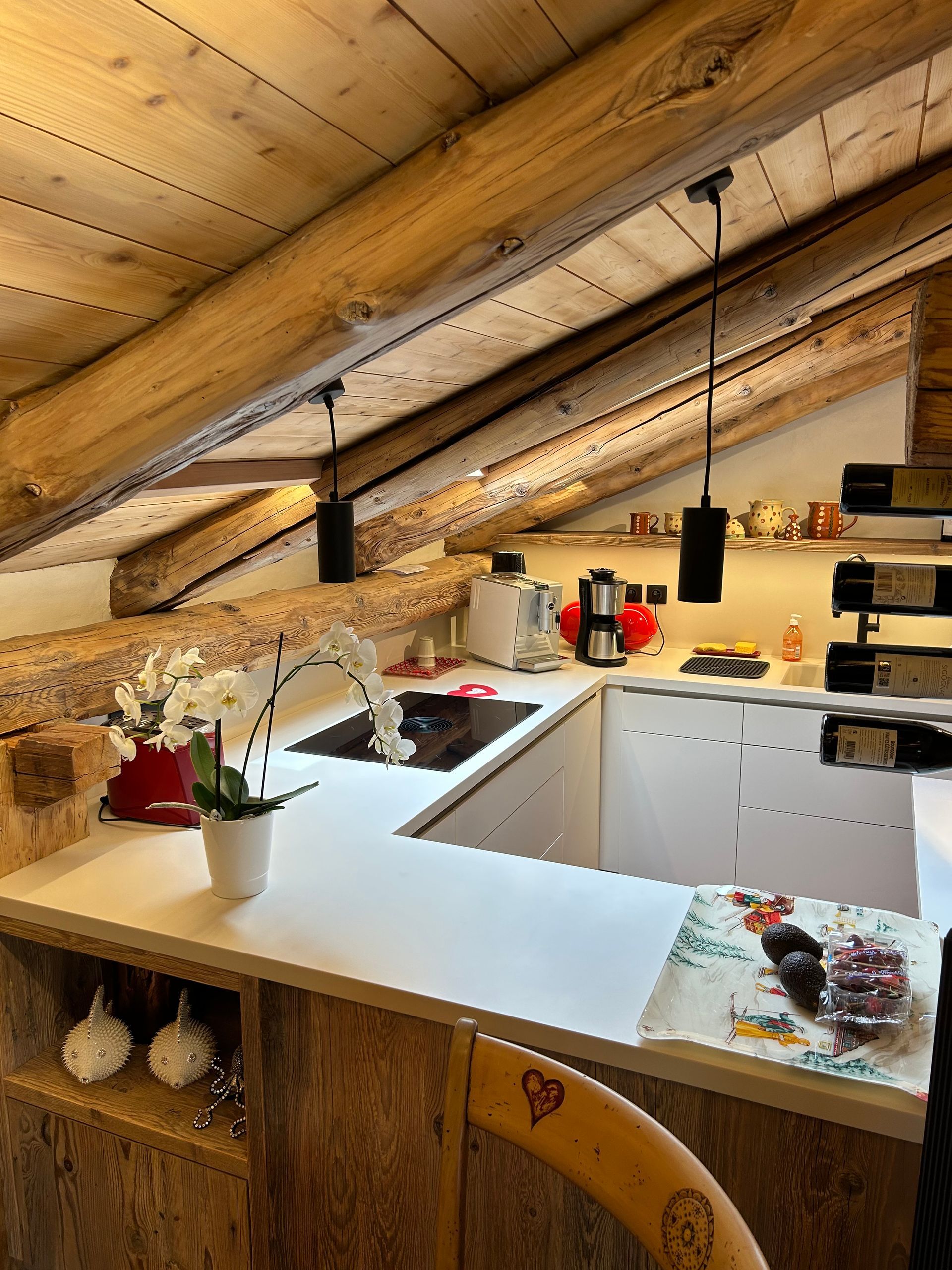 Cuisine chaleureuse avec poutres apparentes et plans de travail blancs. Plaque de cuisson, évier et éclairage au plafond.