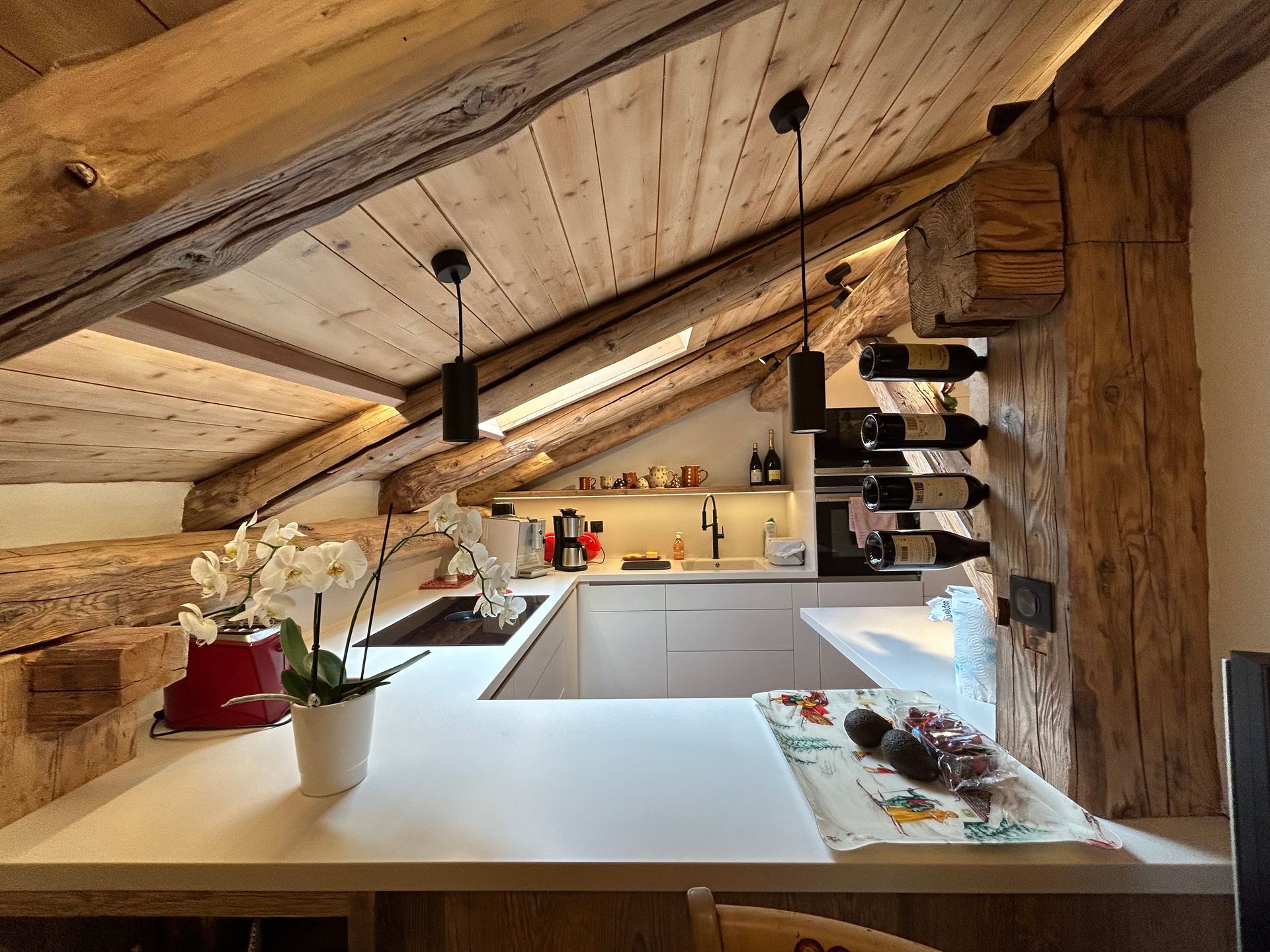 Cuisine mansardée chaleureuse avec poutres apparentes, plans de travail blancs et casier à vin intégré.