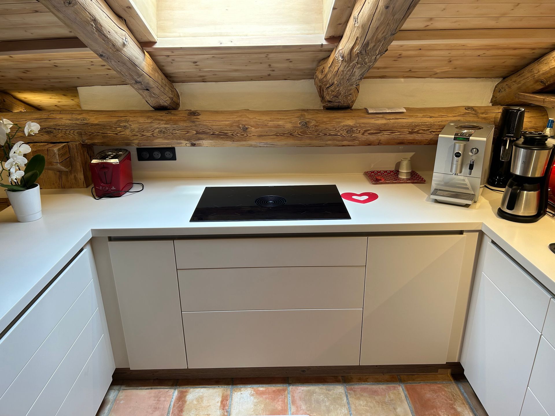 Cuisine blanche avec poutres apparentes et plaque de cuisson intégrée. Les placards sont blancs avec des poignées argentées.