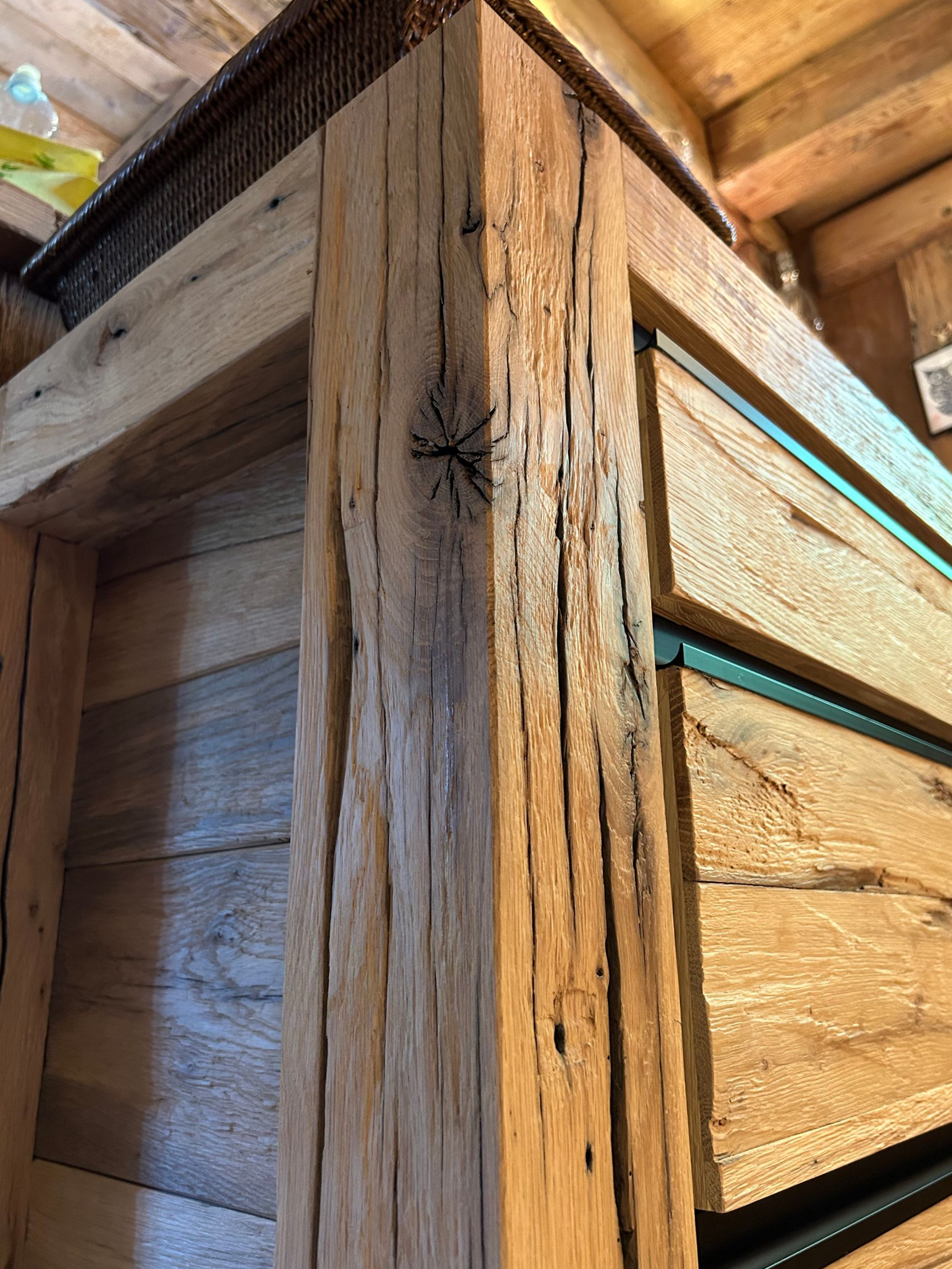 Coin d'un meuble en bois aux poutres rustiques et patinées, laissant apparaître des tiroirs et un panier tressé sur le dessus.