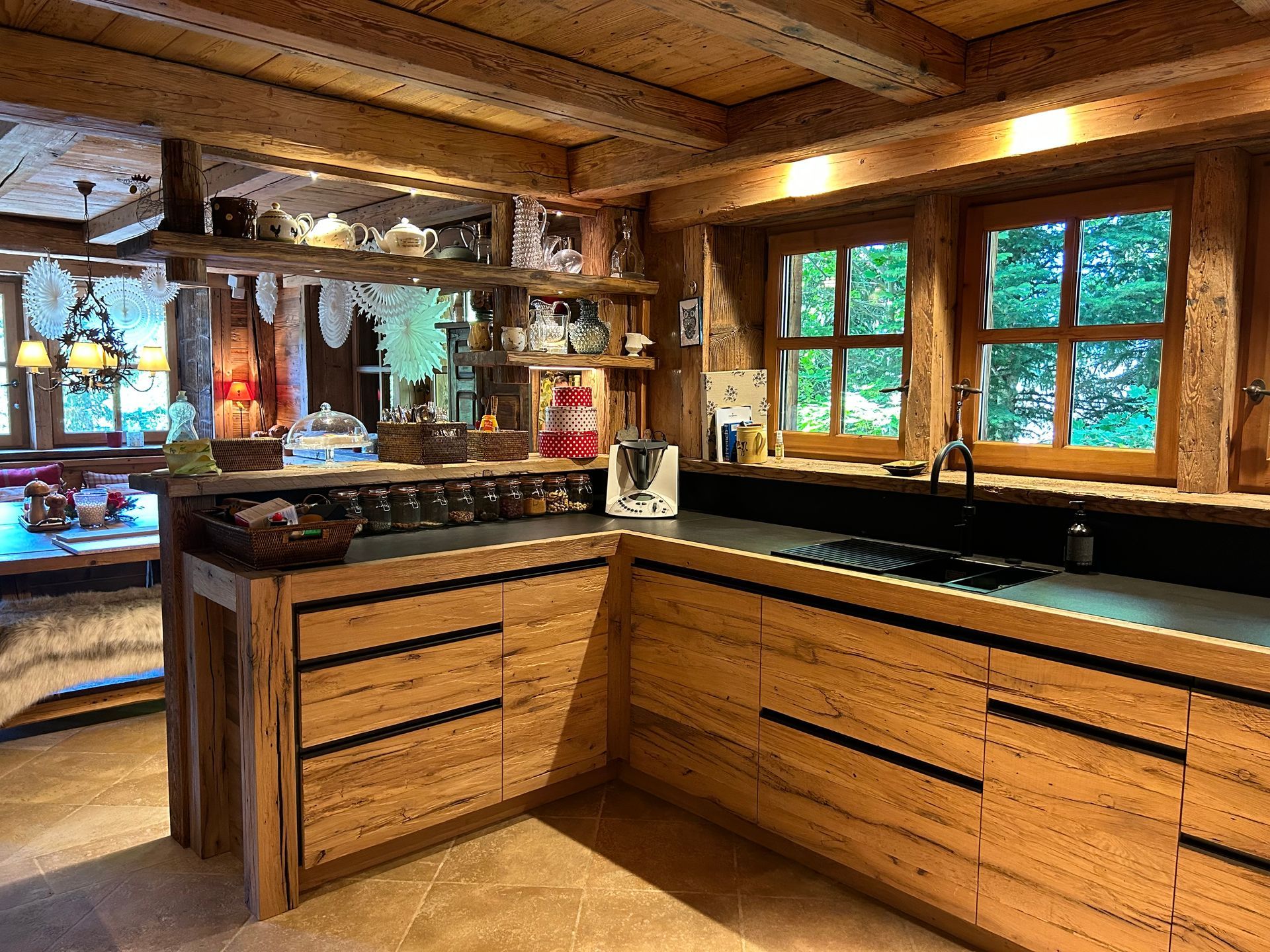 Cuisine en bois avec fenêtres, placards et étagères ouvertes remplies d'objets.