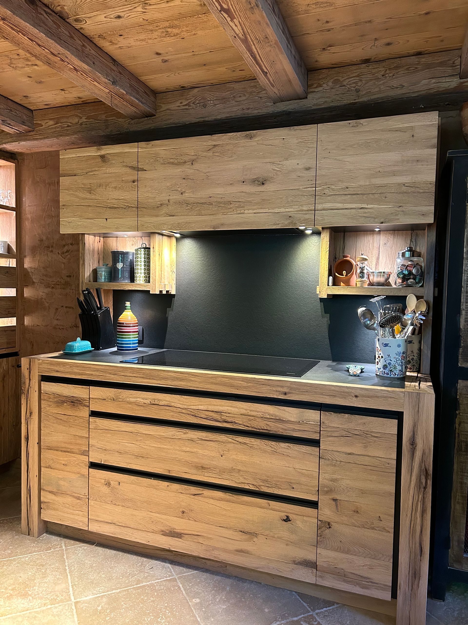 Cuisine équipée d'éléments en bois, d'une cuisinière et d'une crédence noire. Des étagères permettent de ranger bouteilles et autres objets.