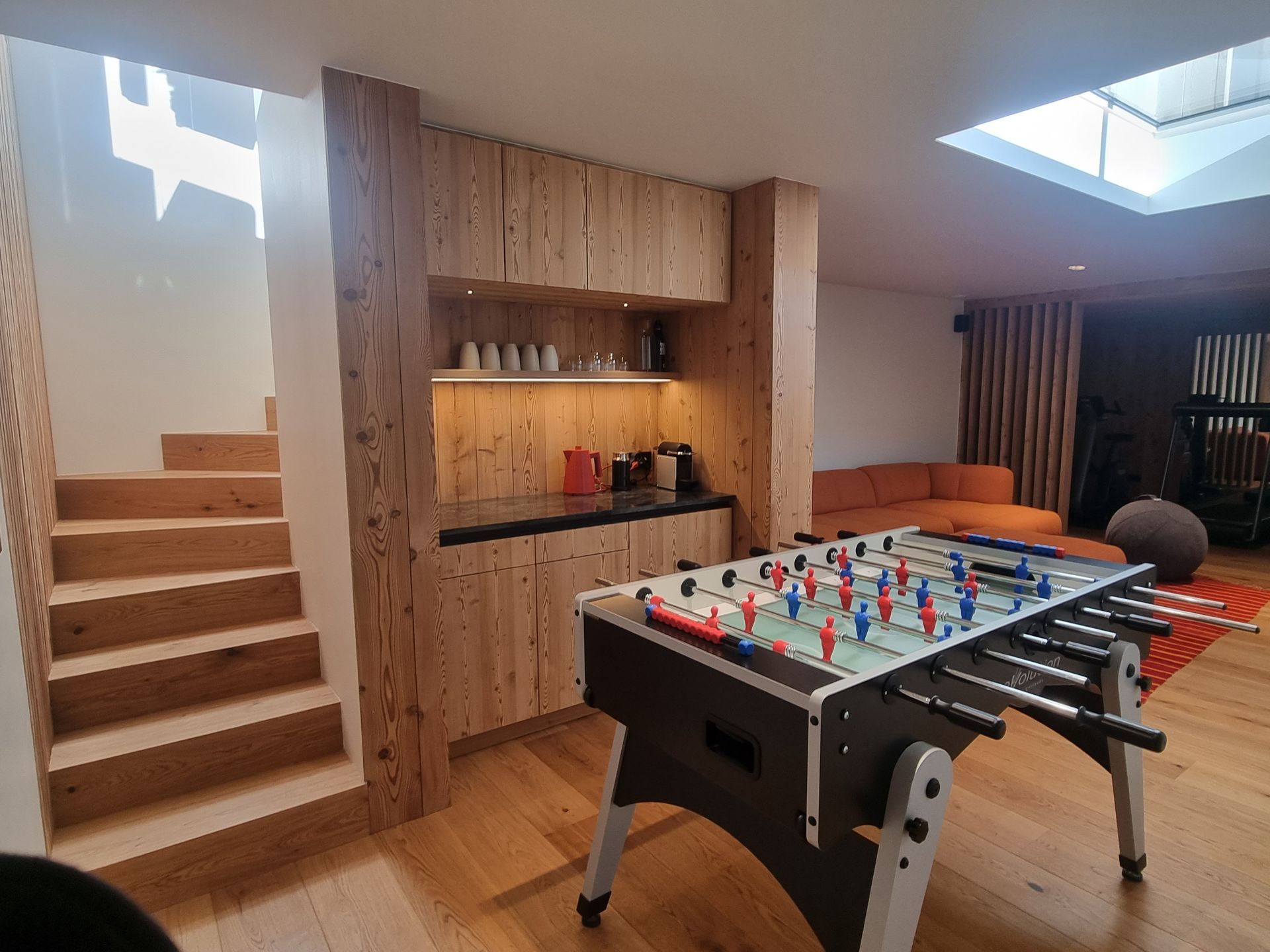 Salle de jeux lambrissée avec baby-foot, escalier et coin cuisine. Canapé orange en arrière-plan, puits de lumière au-dessus.