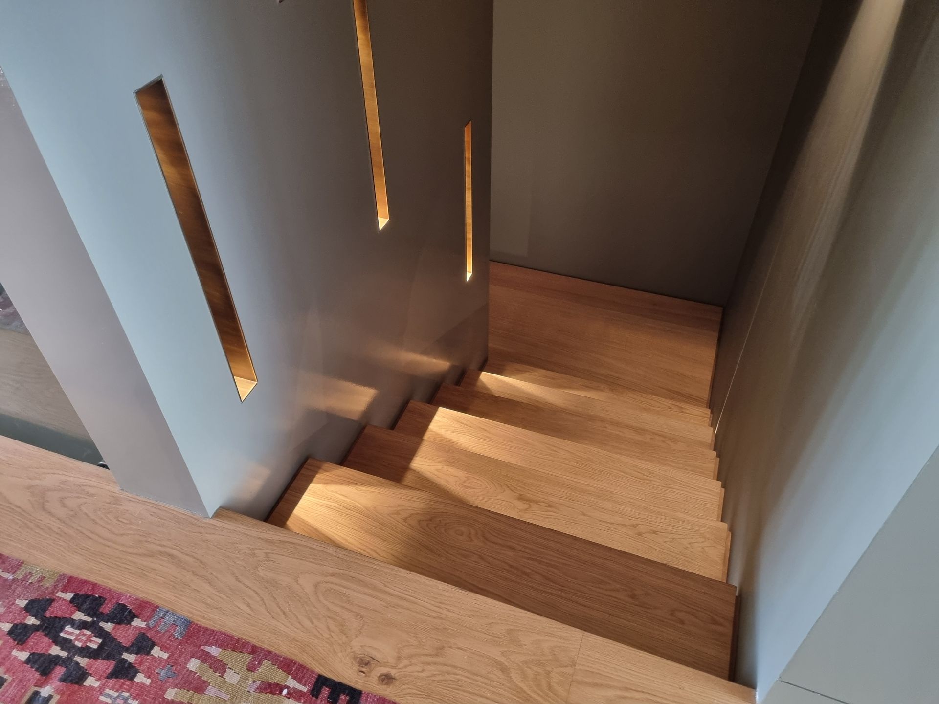 Un escalier en bois descend dans un coin, éclairé par des ouvertures dans le mur. Murs gris, escalier en bois clair.