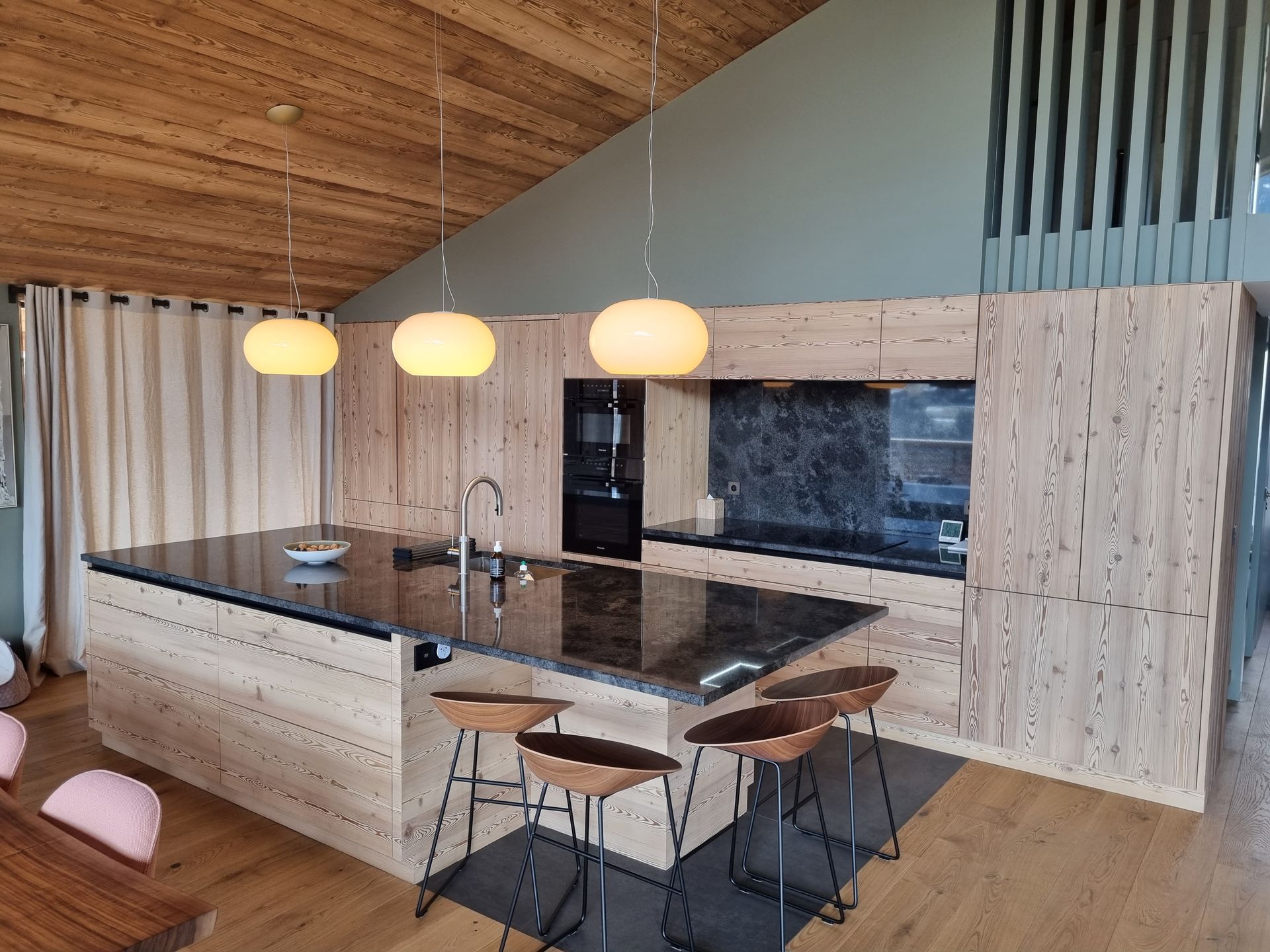 Cuisine moderne avec placards en bois clair, plans de travail noirs et trois suspensions lumineuses.