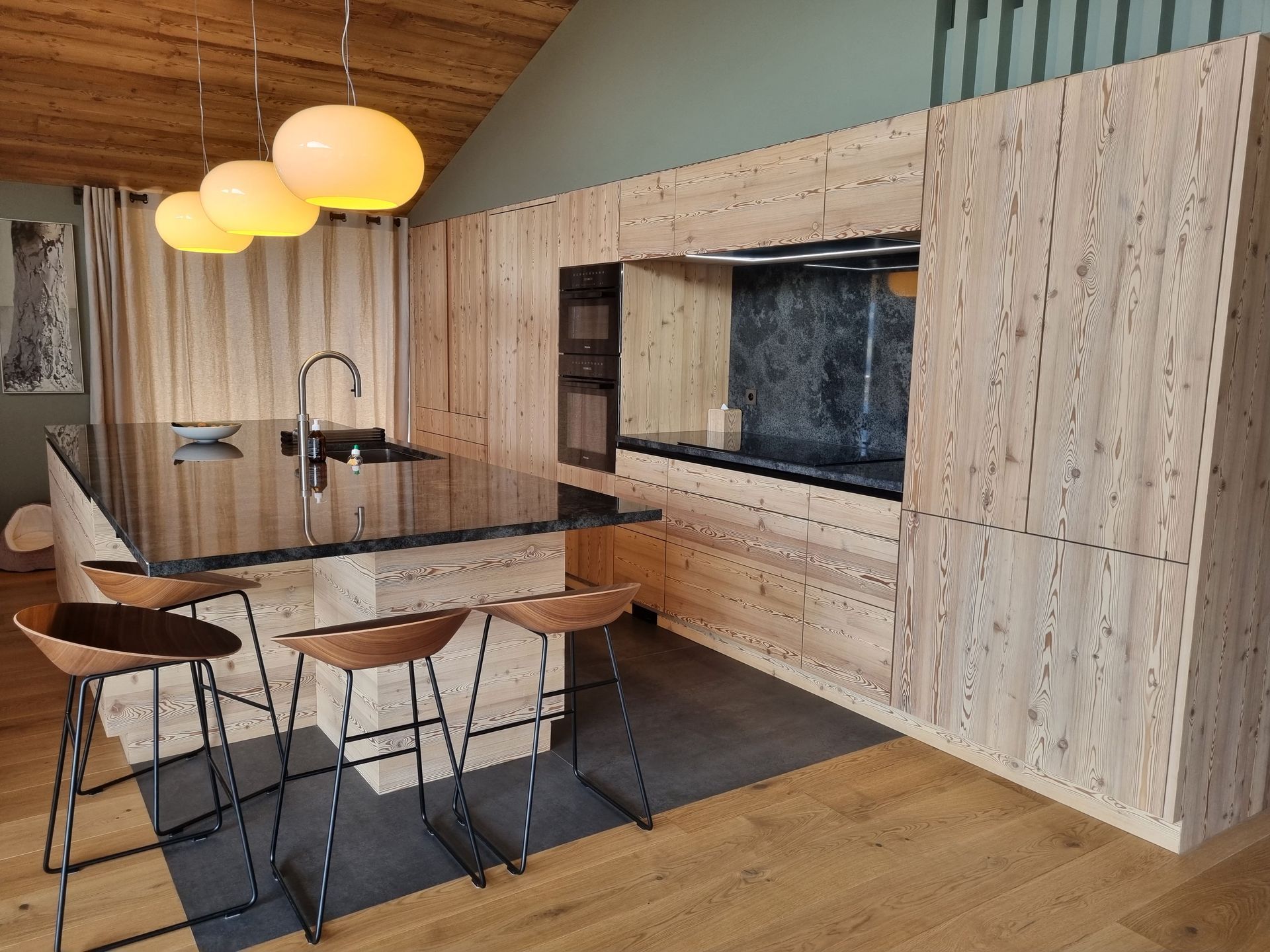 Cuisine moderne avec placards en bois clair, plans de travail noirs et suspensions lumineuses.