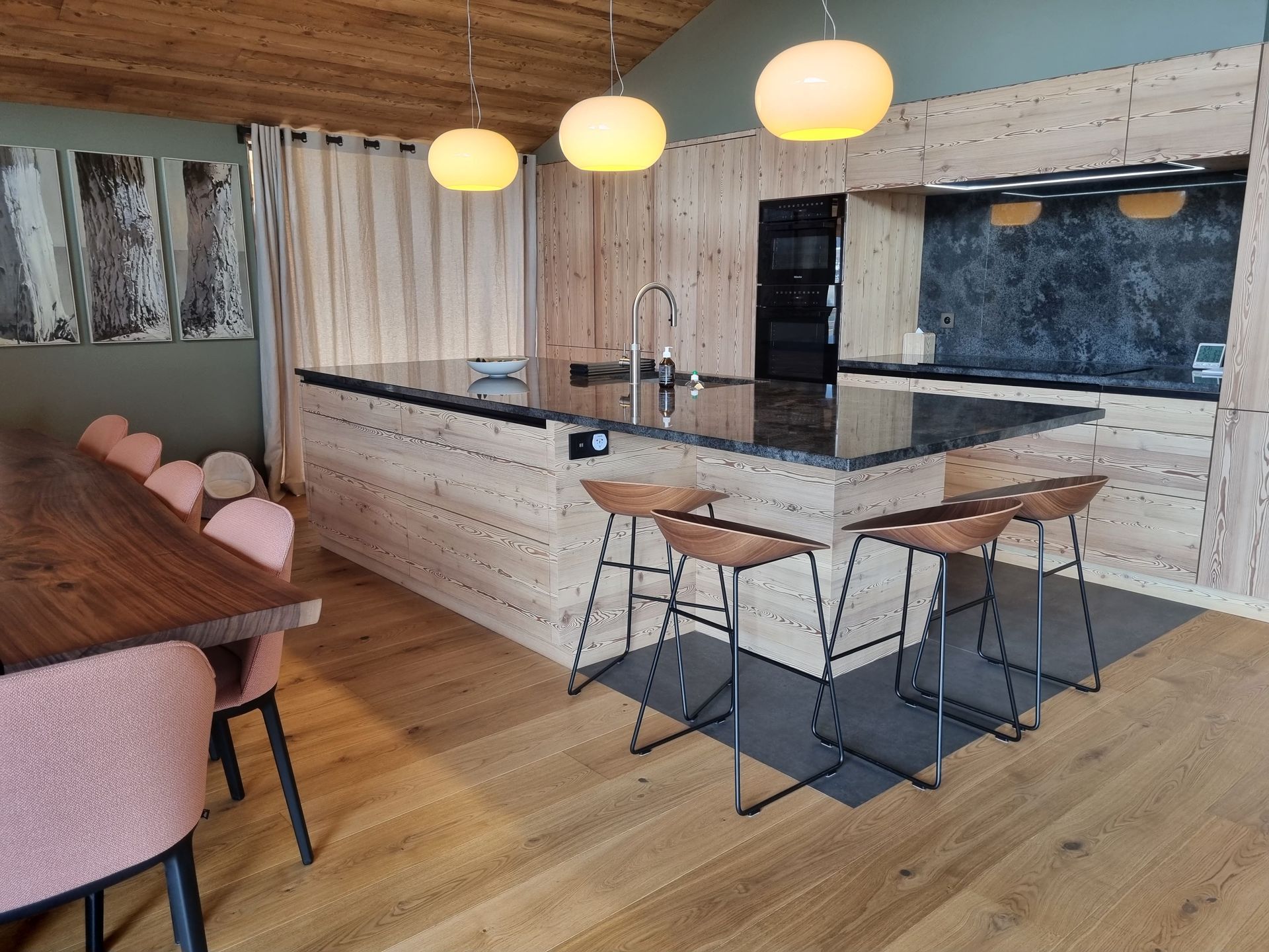 Cuisine en bois avec îlot central, tabourets et suspensions lumineuses. Table à manger avec chaises roses à gauche.