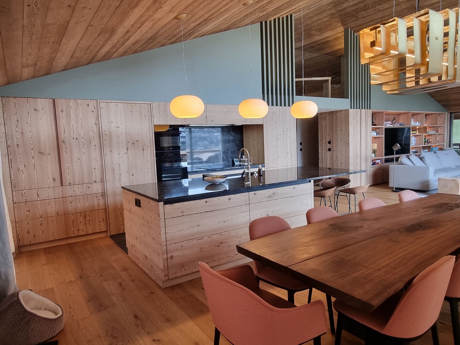 Cuisine avec placards en bois, îlot central et table à manger ; trois suspensions lumineuses surplombent l'îlot.