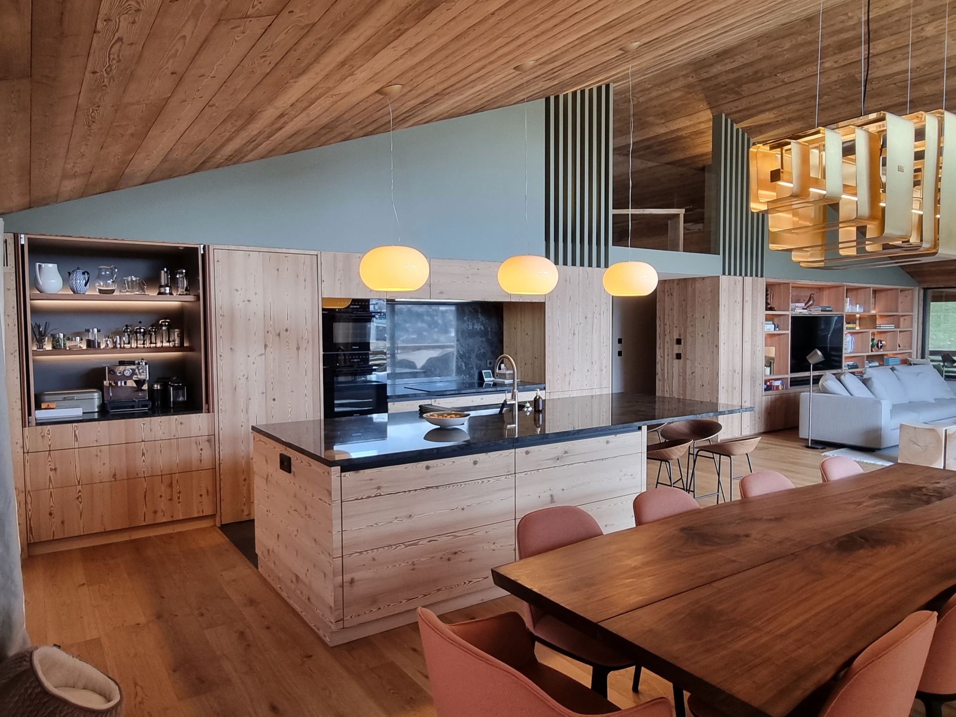 Cuisine en bois avec îlot central et table à manger, luminaires suspendus et étagères intégrées.