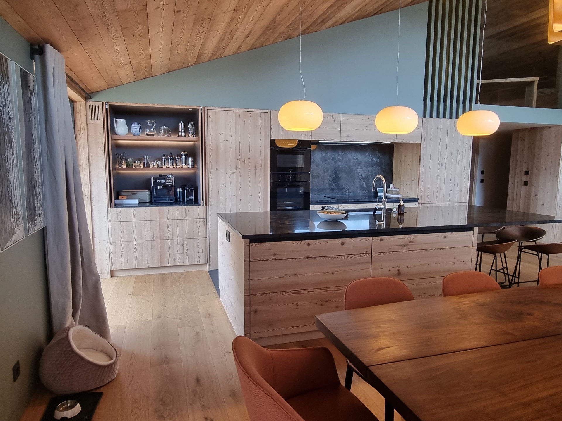 Cuisine en bois avec îlot central, placards, four et suspensions lumineuses. Table à manger avec chaises orange.