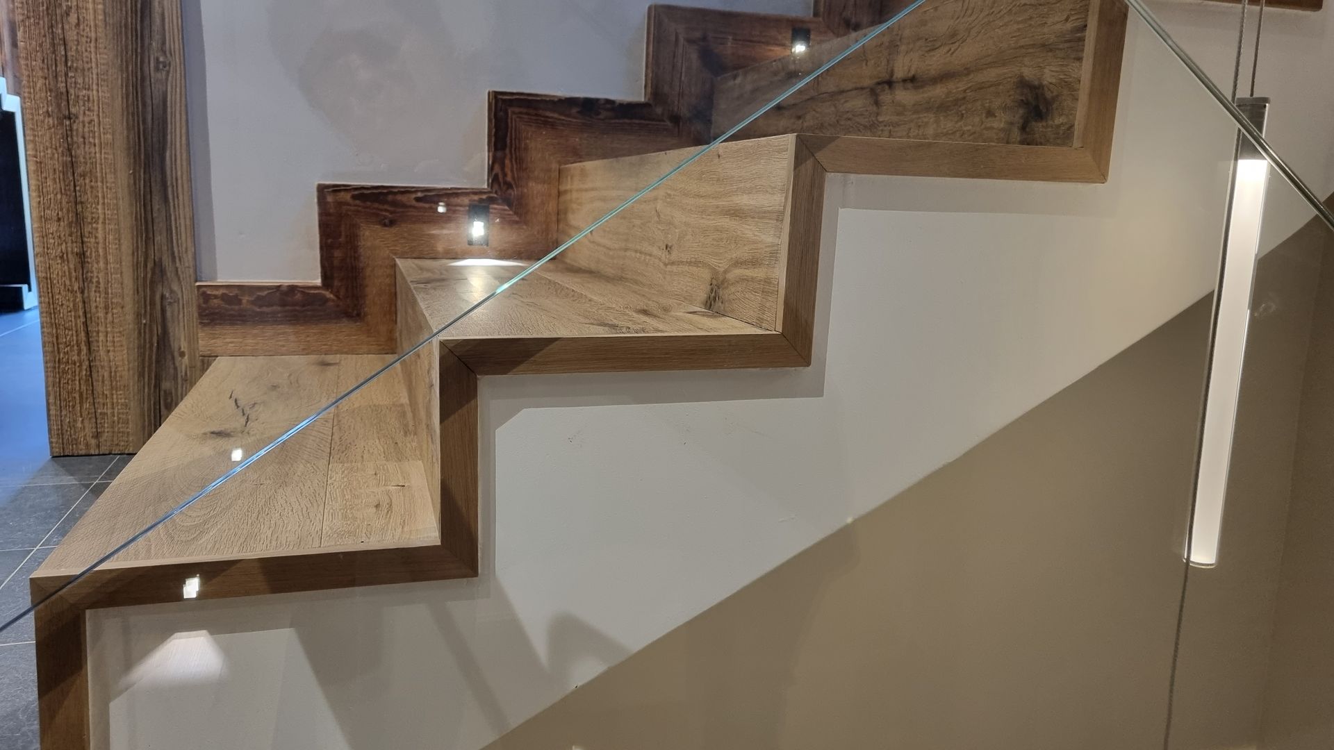 Escalier en bois avec éclairage intégré, rampe en verre, murs beiges et marches en pierre brune.