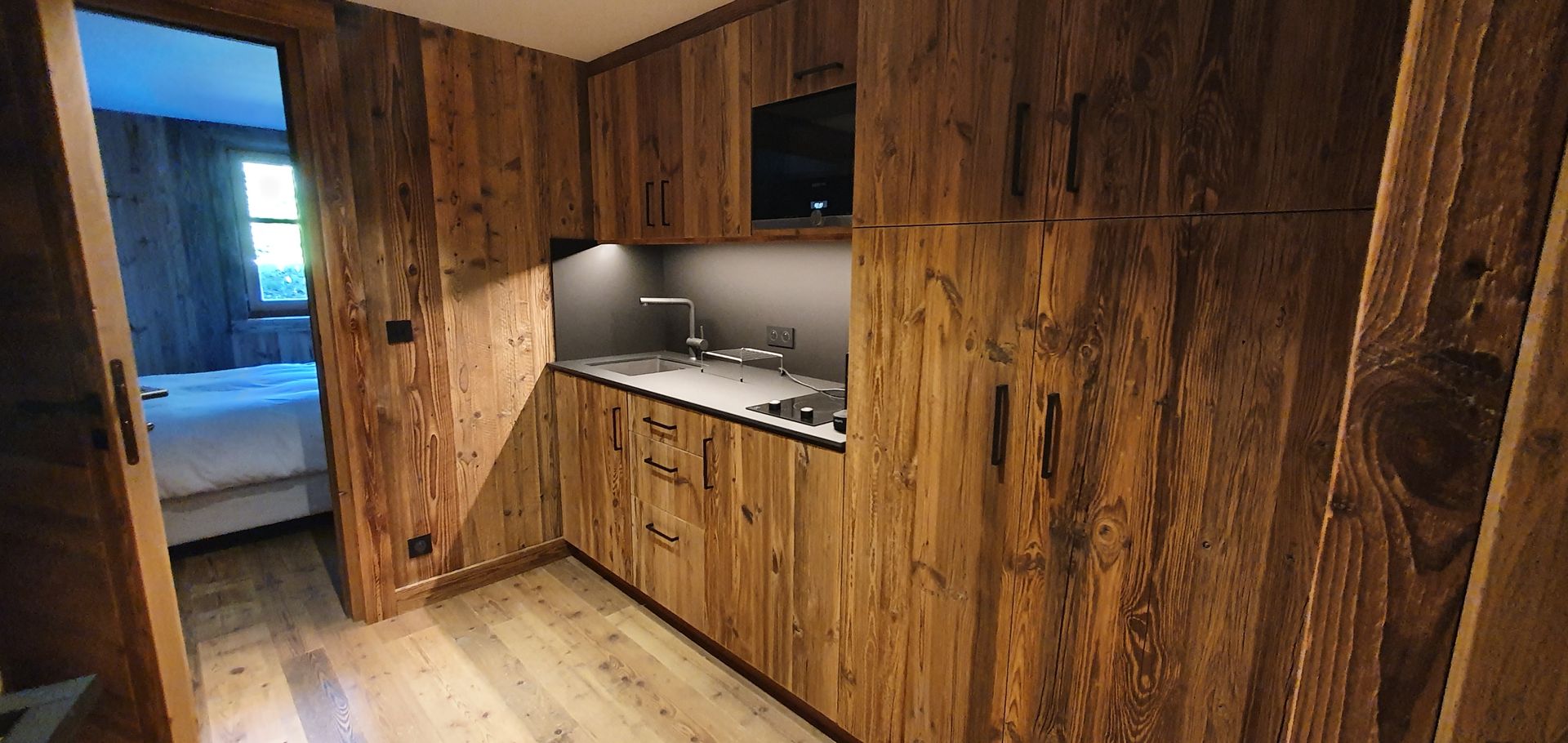 Petite cuisine avec placards en bois, plan de travail et électroménager. Porte visible à droite.