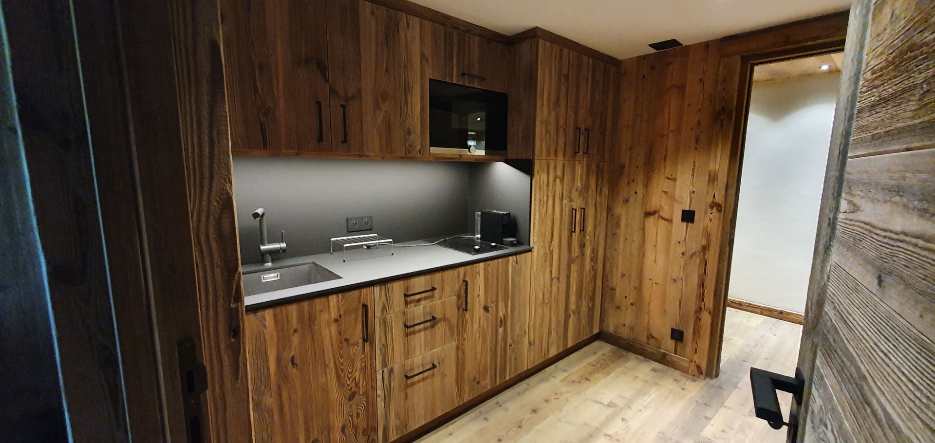 Petite cuisine en bois avec placards, évier, micro-ondes et porte ouverte.