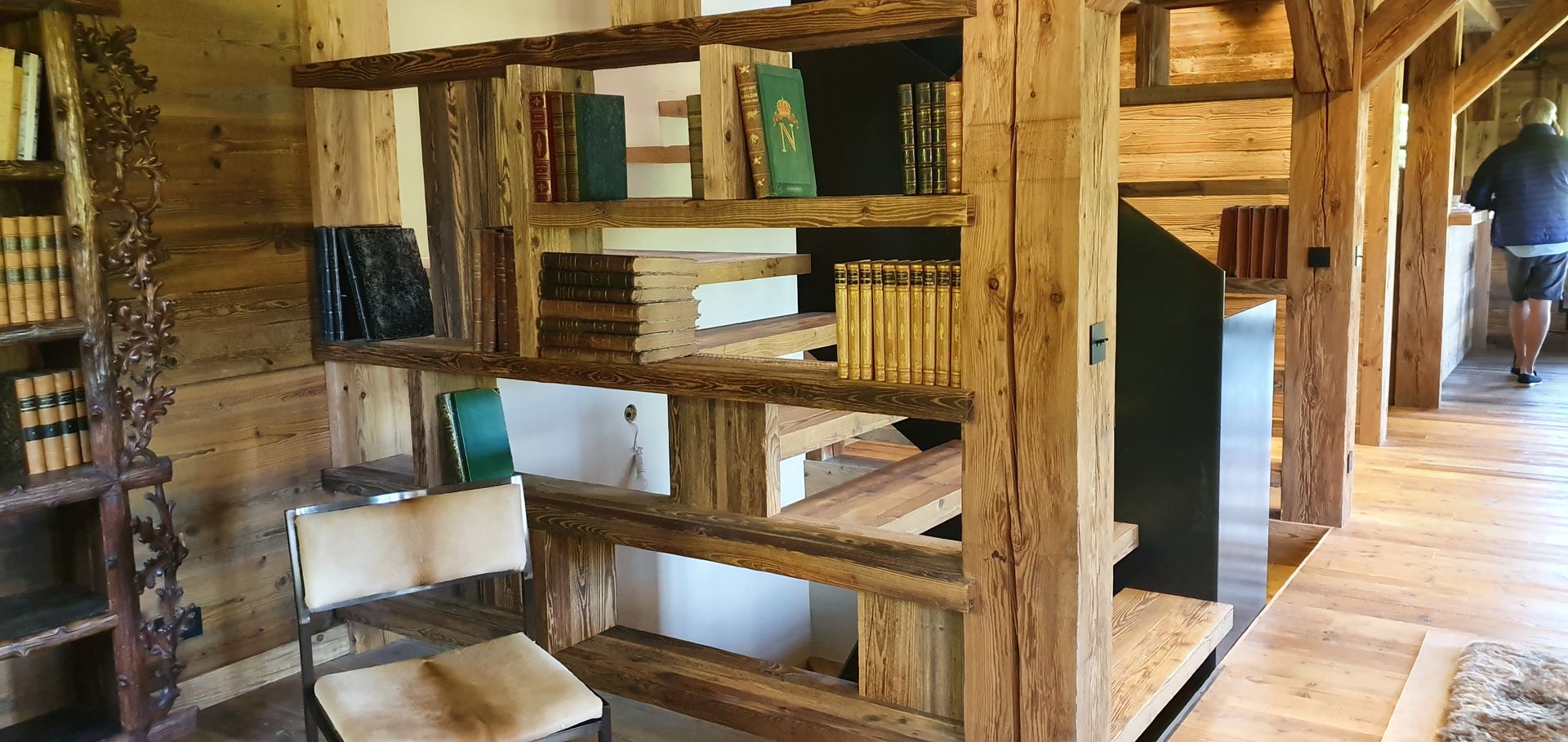 Des étagères en bois remplies de livres. Une chaise est placée devant. Une personne marche à l'arrière-plan.